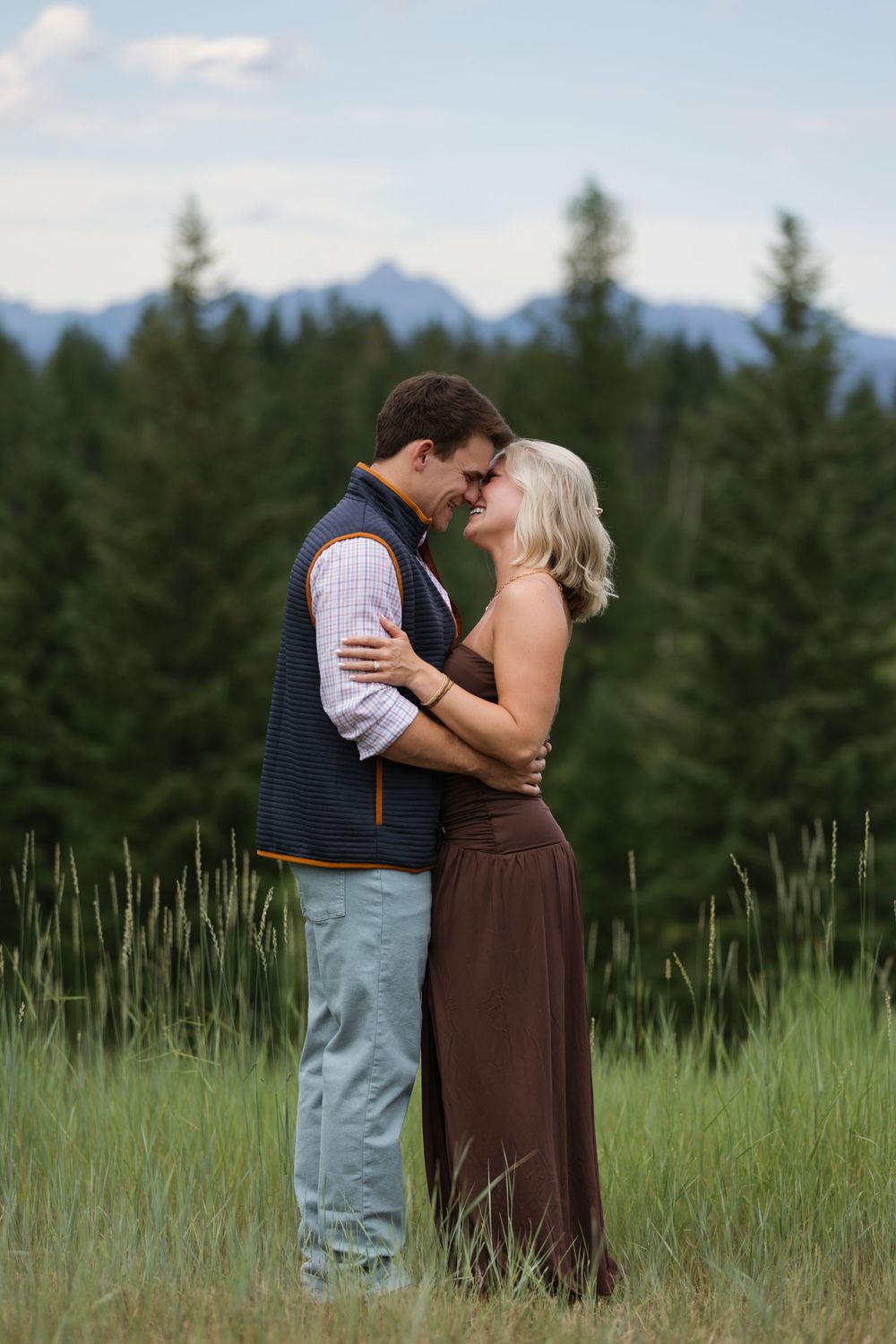 Couple embracing in a lush green meadow with pine trees and mountains in the background.