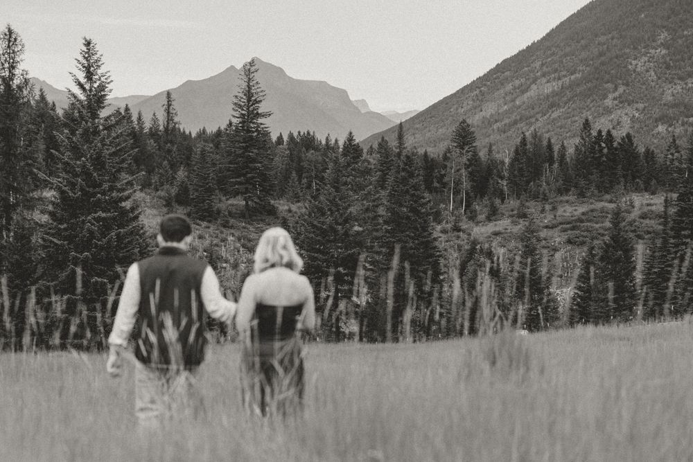 Black and white photo of a couple holding hands walking through a mountain meadow with pine trees and peaks behind them.