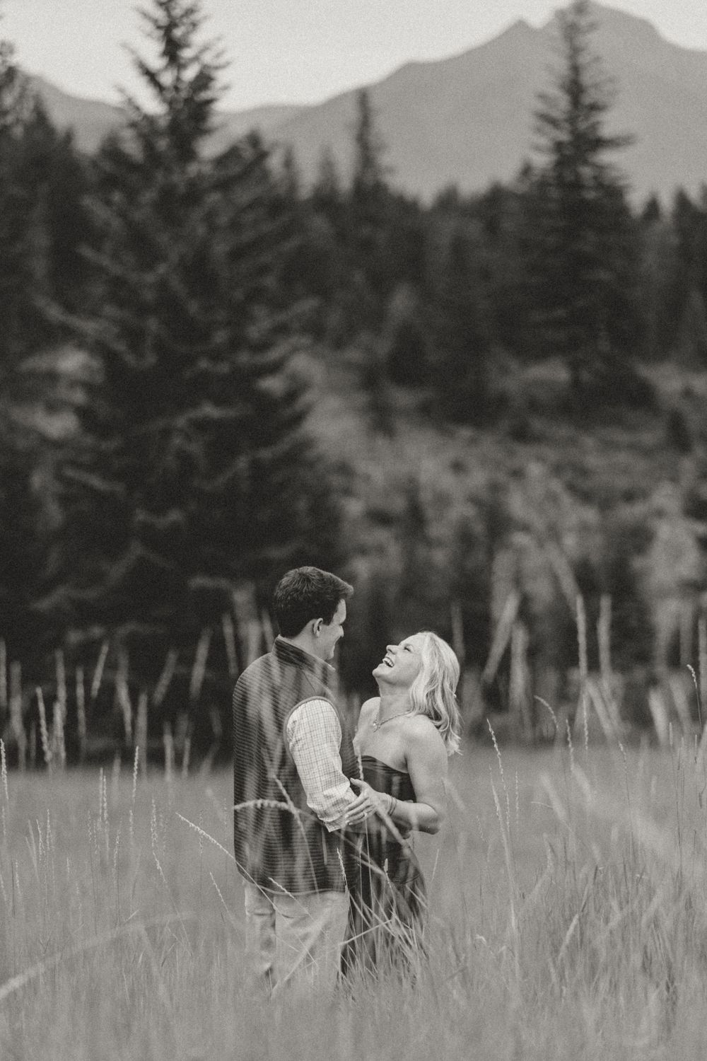 Black and white photo of a couple laughing together in a mountain meadow surrounded by tall grass and pine trees.