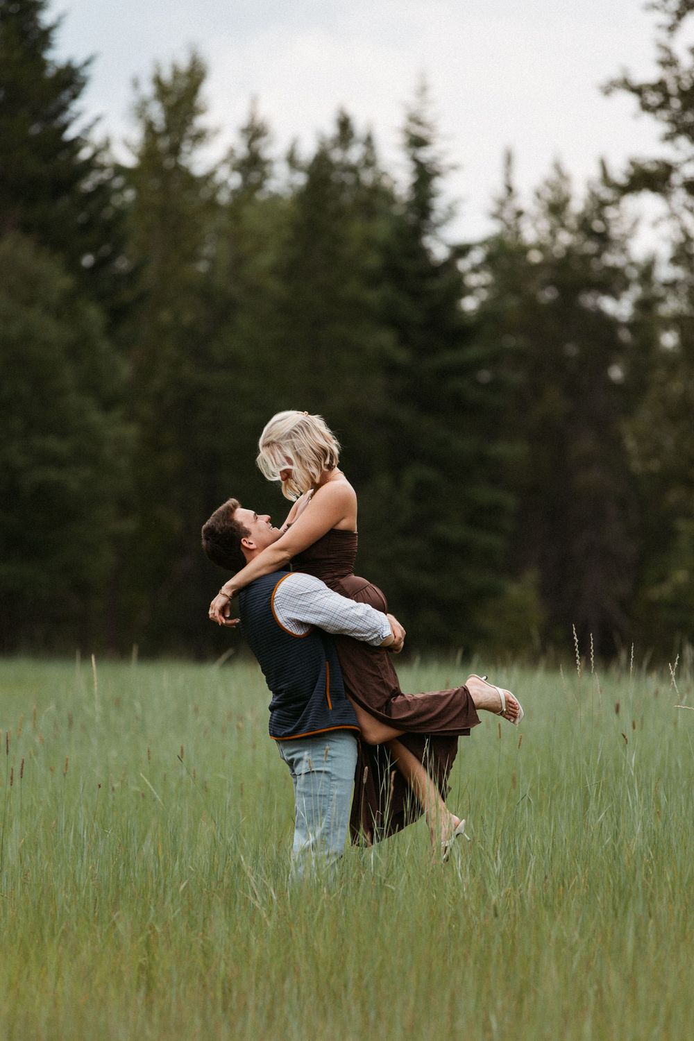 A man lifts a woman in a green meadow, sharing a romantic moment with tall pine trees in the background.