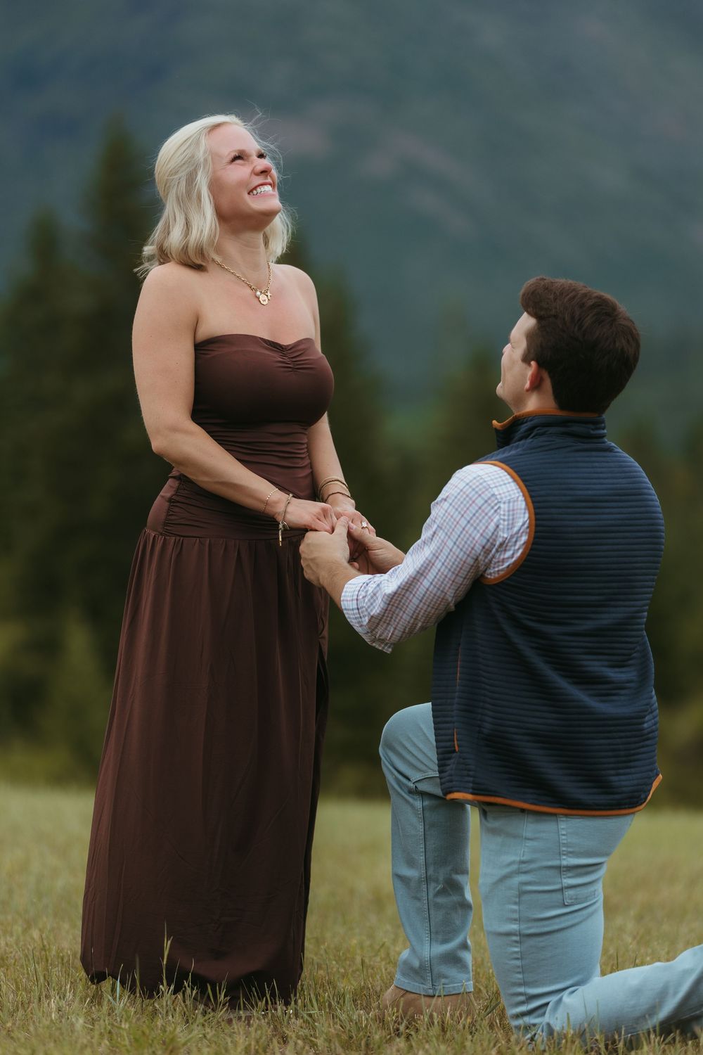 Man kneeling on one knee proposing to woman in brown dress outdoors with mountain backdrop.