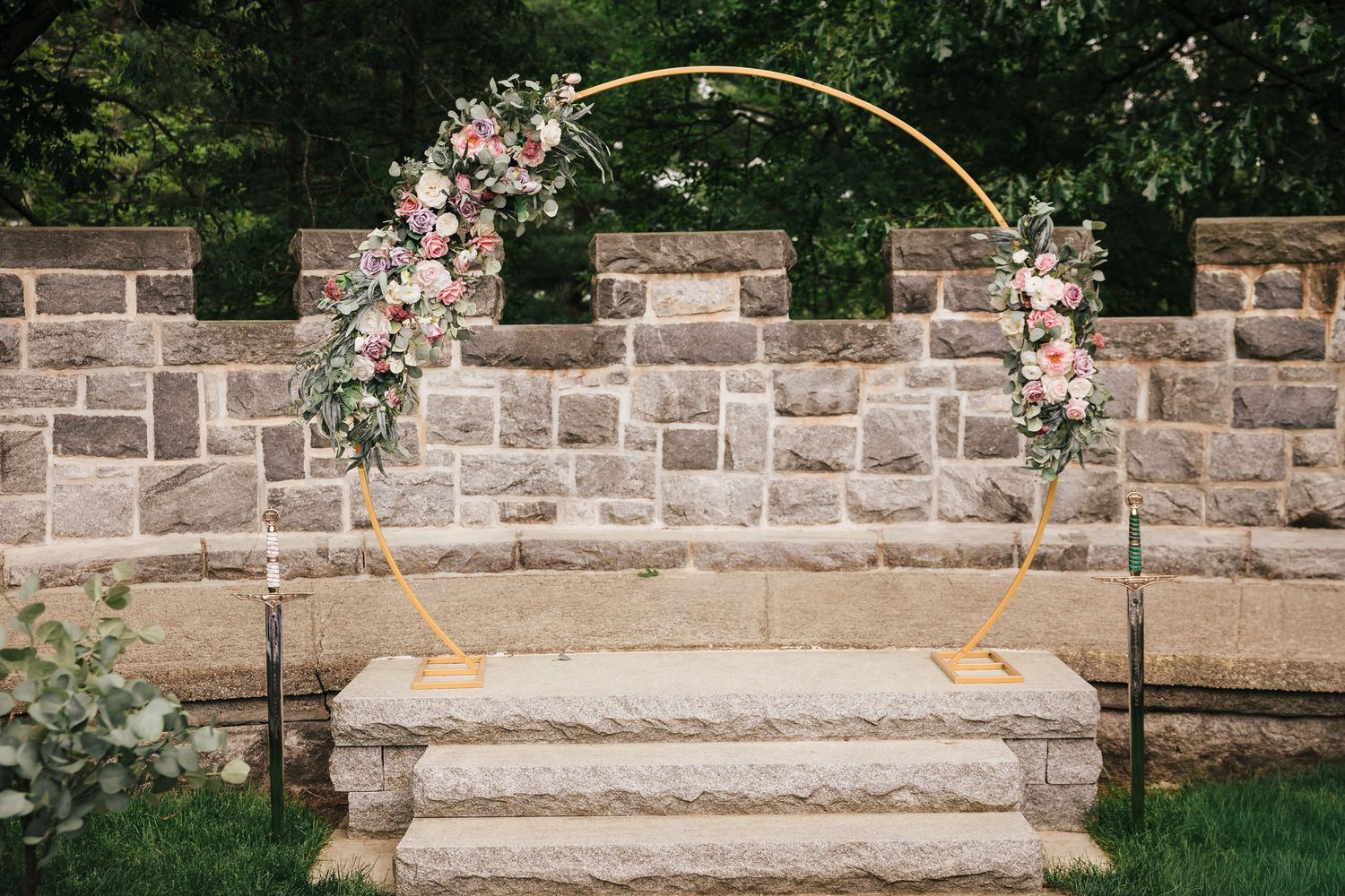 Gold ceremony arch with pink and white floral clusters, flanked by two ornamental broadswords, at Searles Castle.