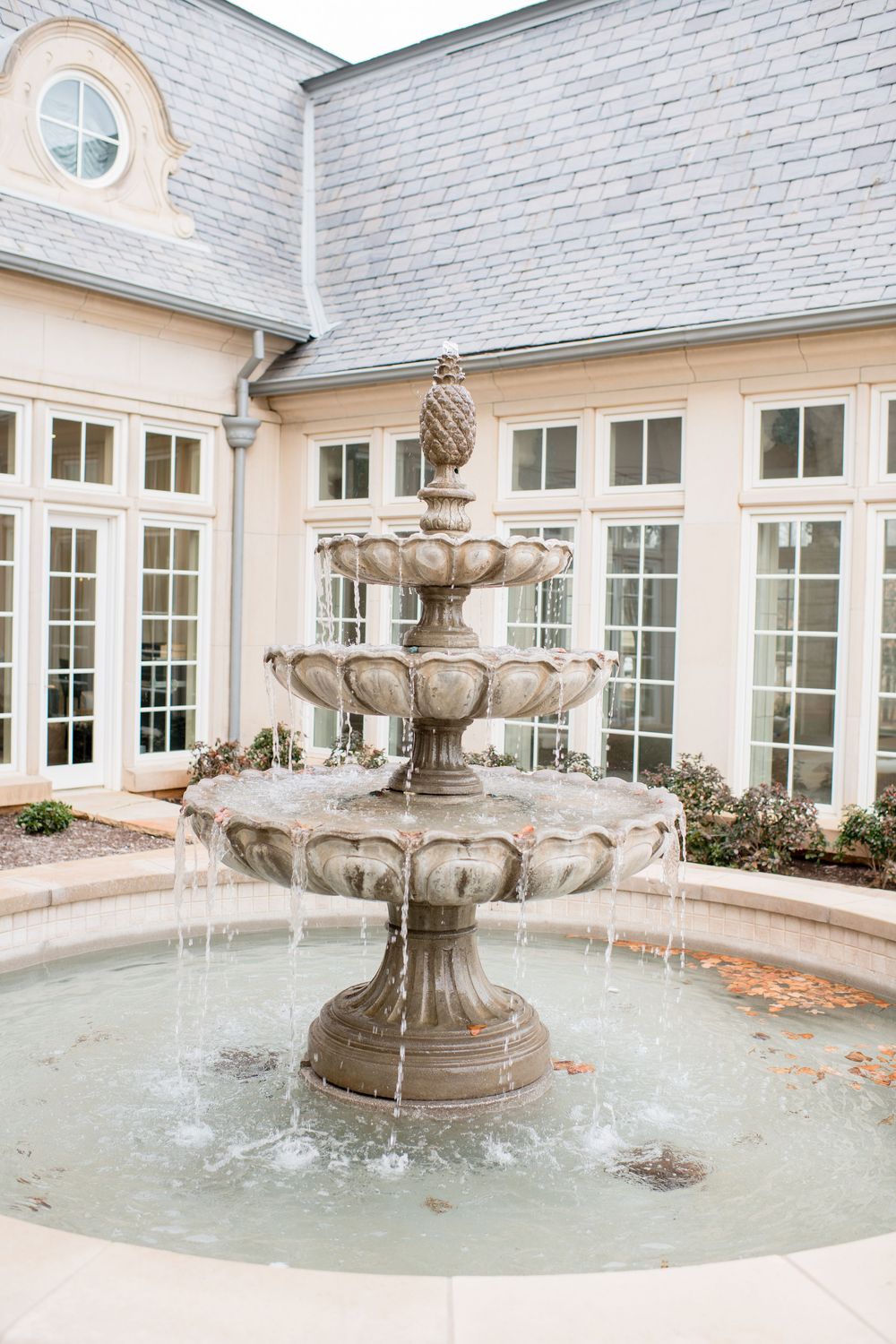 Elegant three-tier stone fountain with pineapple finial in front of a classic white building with French windows.