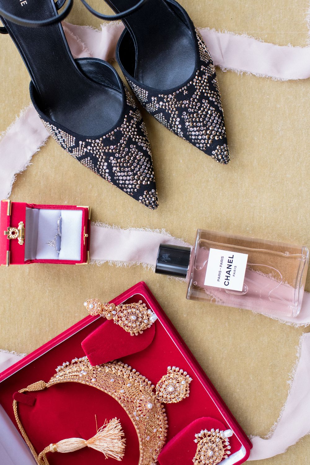 Bridal flat lay with embellished heels, Chanel perfume, diamond ring, and gold Indian jewelry on red velvet tray.