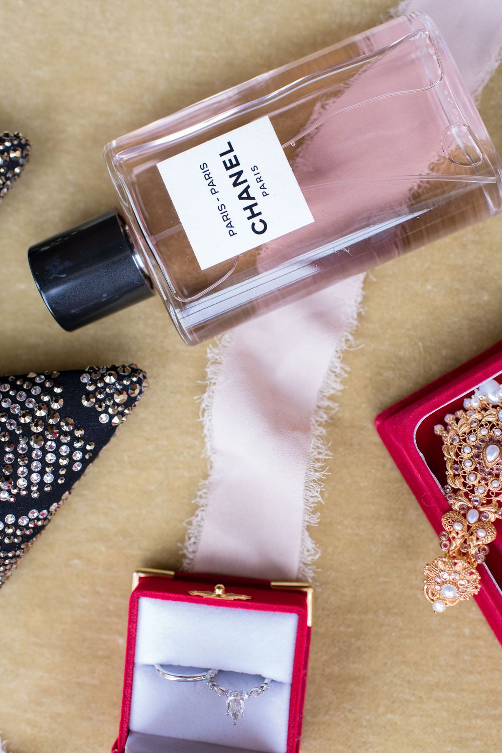 Chanel perfume bottle with pink ribbon, silver ring in red box, rhinestone heels, and gold brooch on beige background.
