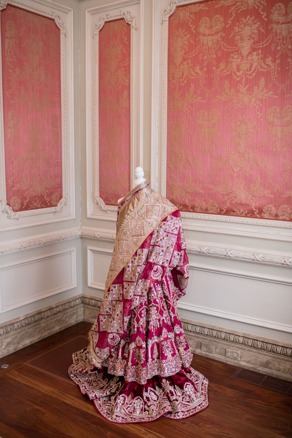 Elegant pink and gold Indian bridal lehenga on mannequin displayed in ornate room with pink damask walls.