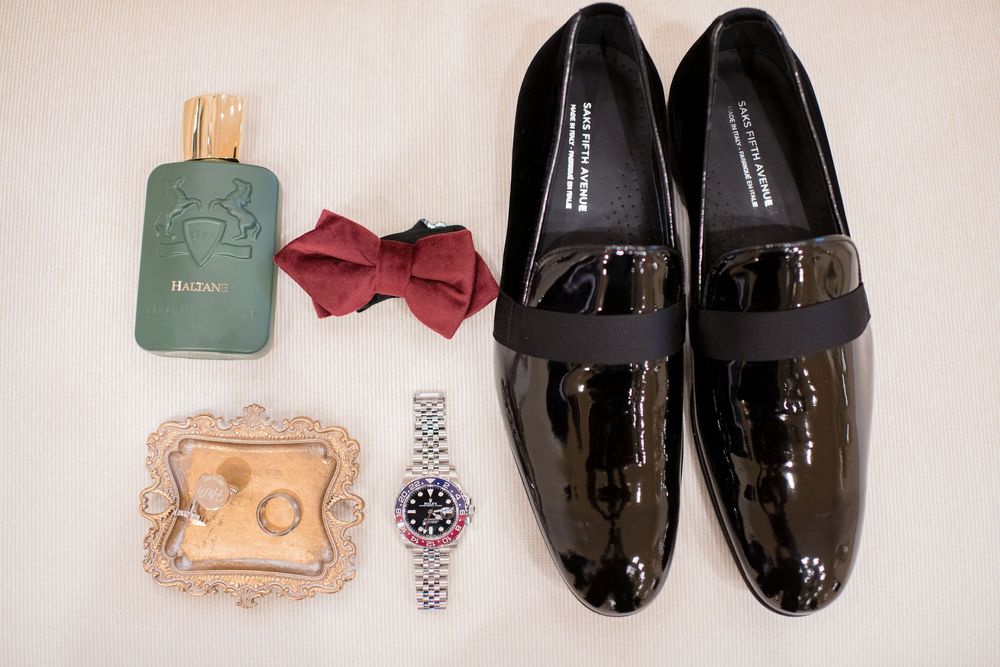 Black patent leather loafers, red bow tie, green cologne bottle, silver Rolex watch, and gold ring tray flat lay.