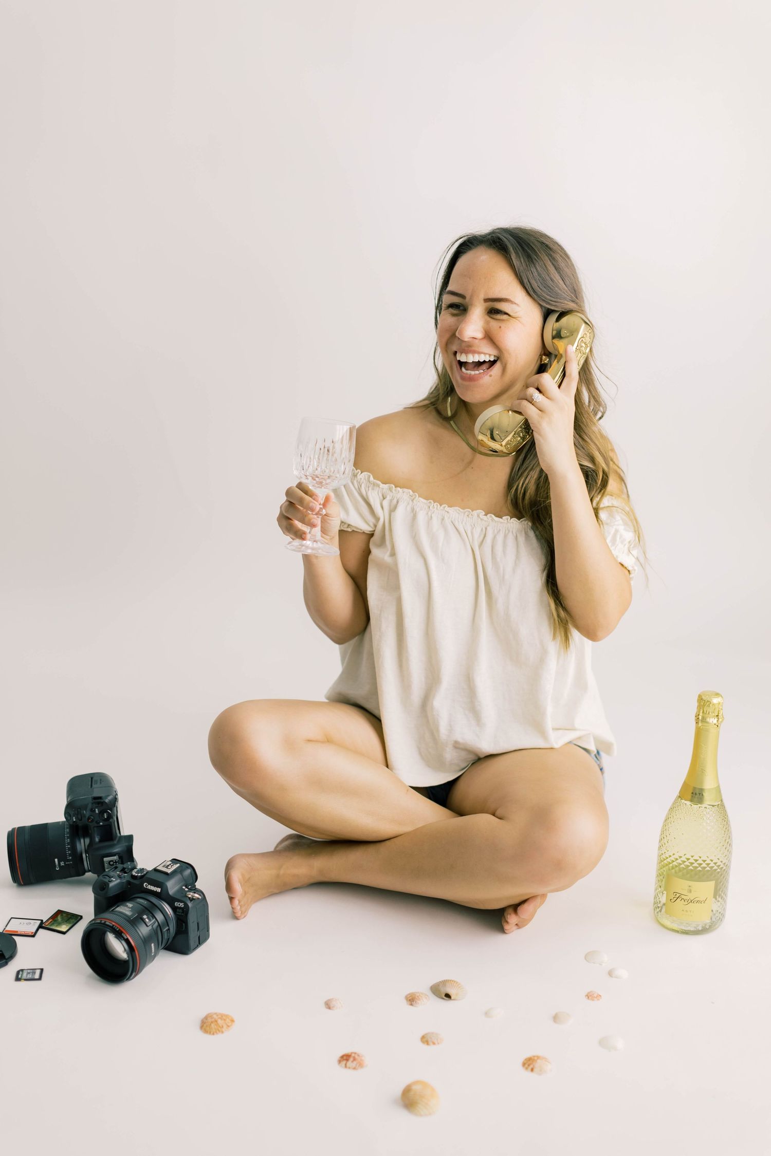 Woman sitting cross-legged holding champagne glass and retro phone, with camera and bottle nearby on white background.