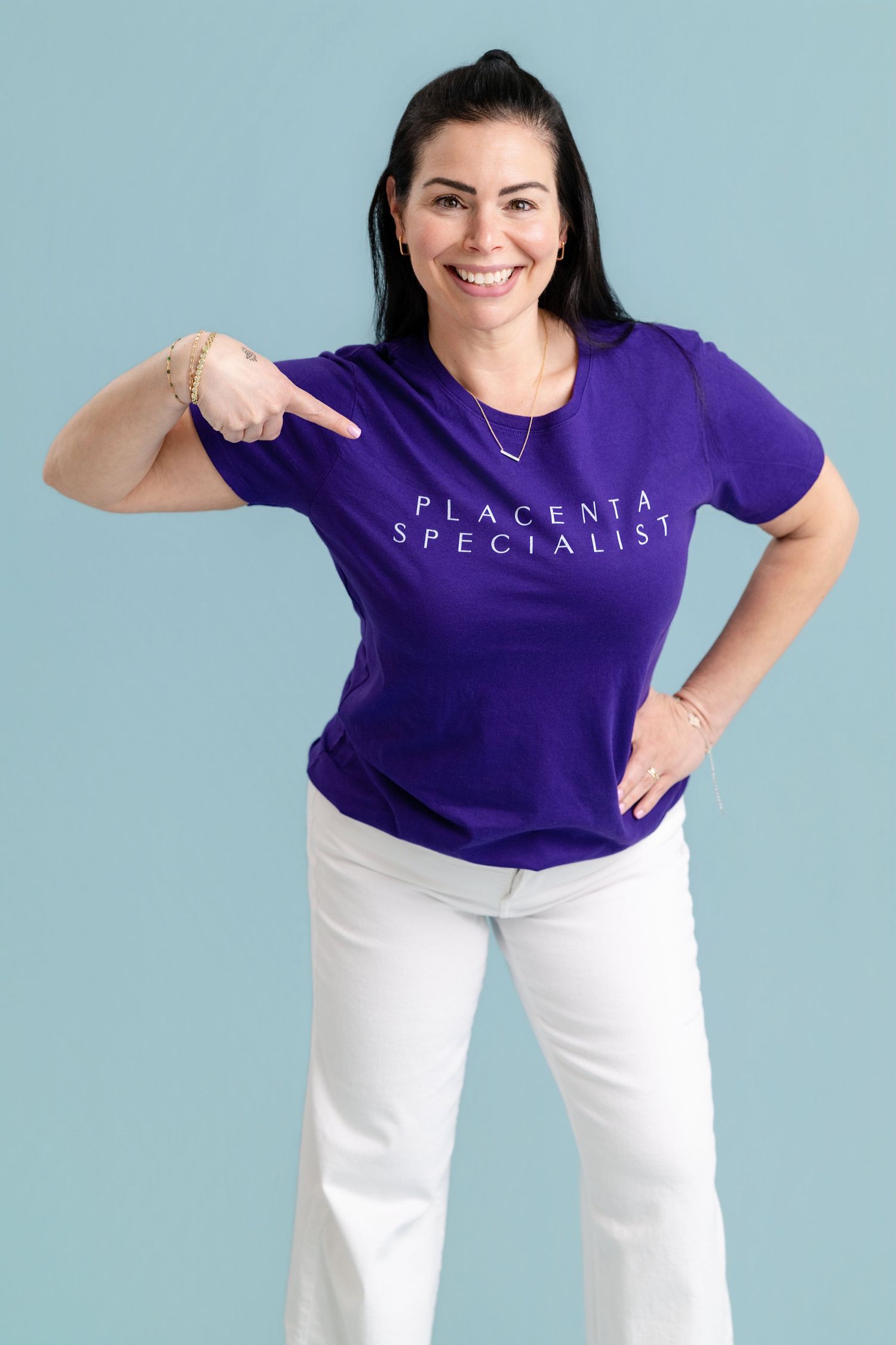 Smiling woman in purple Placenta Specialist t-shirt pointing at text, wearing white jeans on blue background.