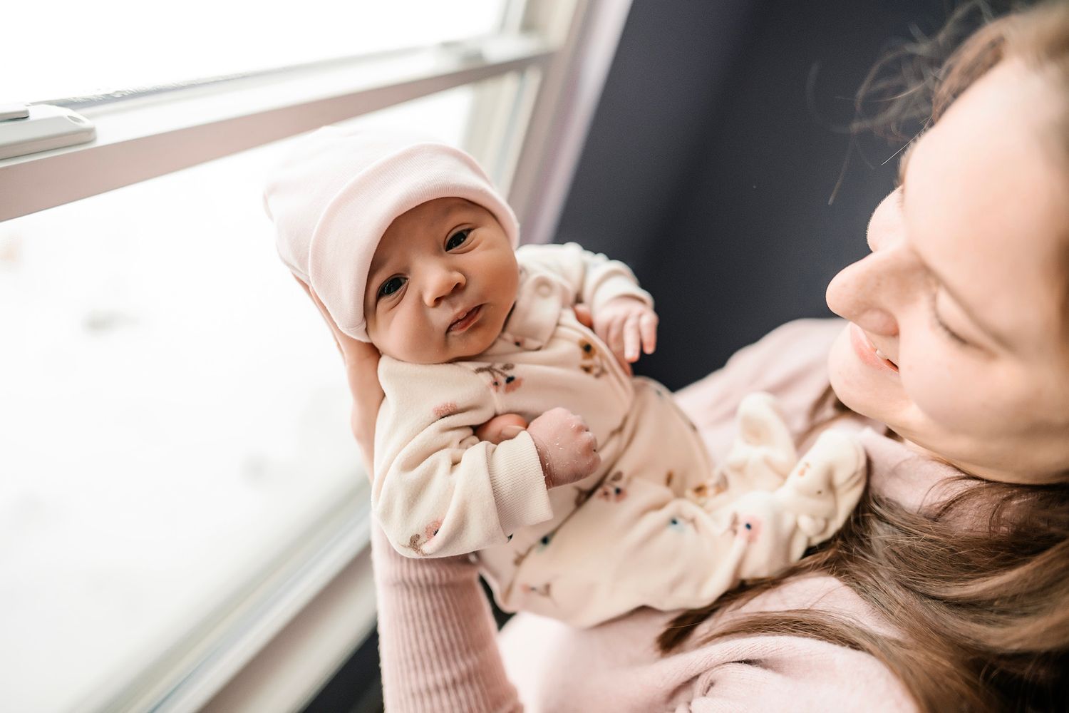 A mother tenderly holds her newborn baby in a pink outfit and hat near a bright window.