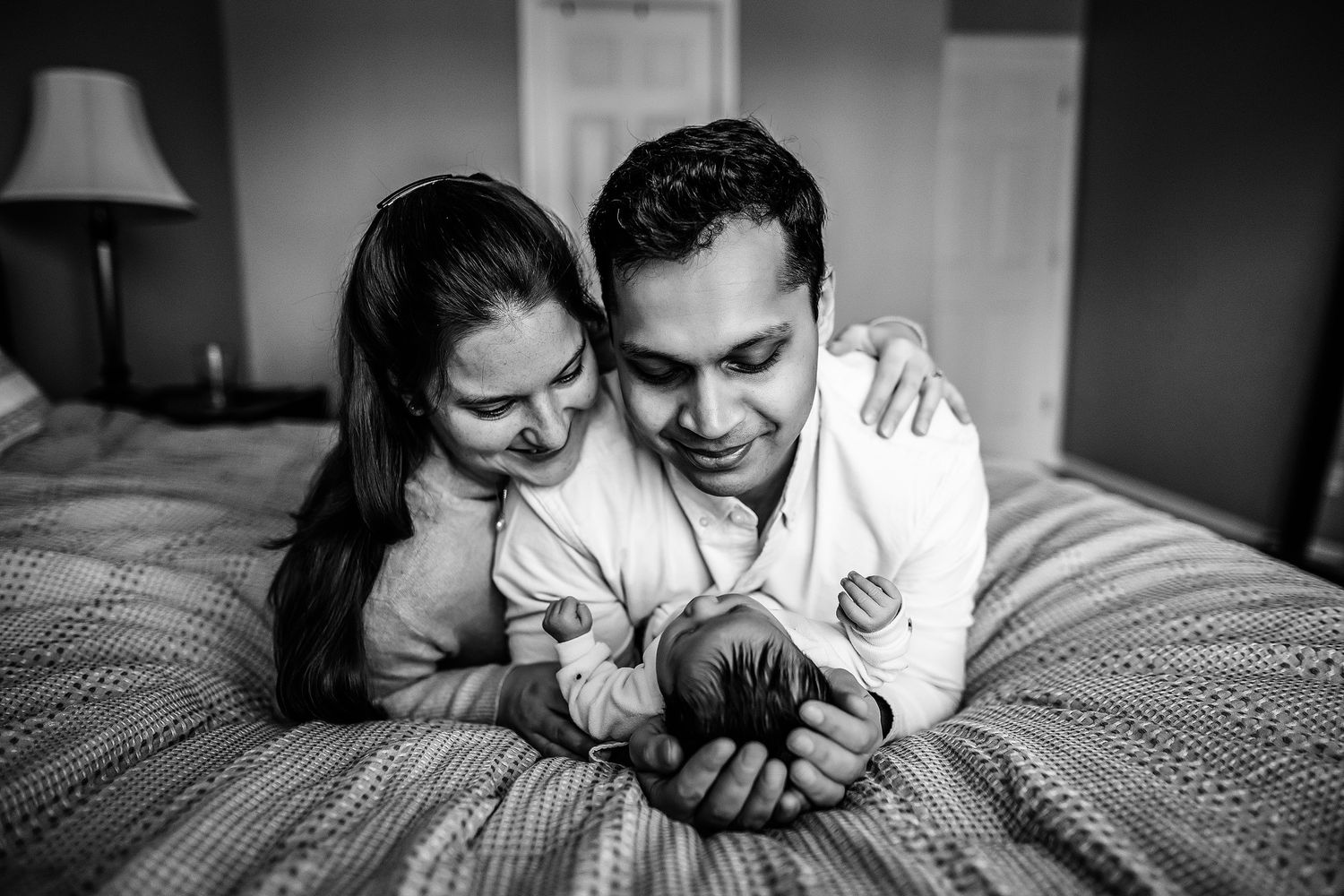 Black and white photo of smiling parents lying on bed gazing lovingly at their newborn baby.