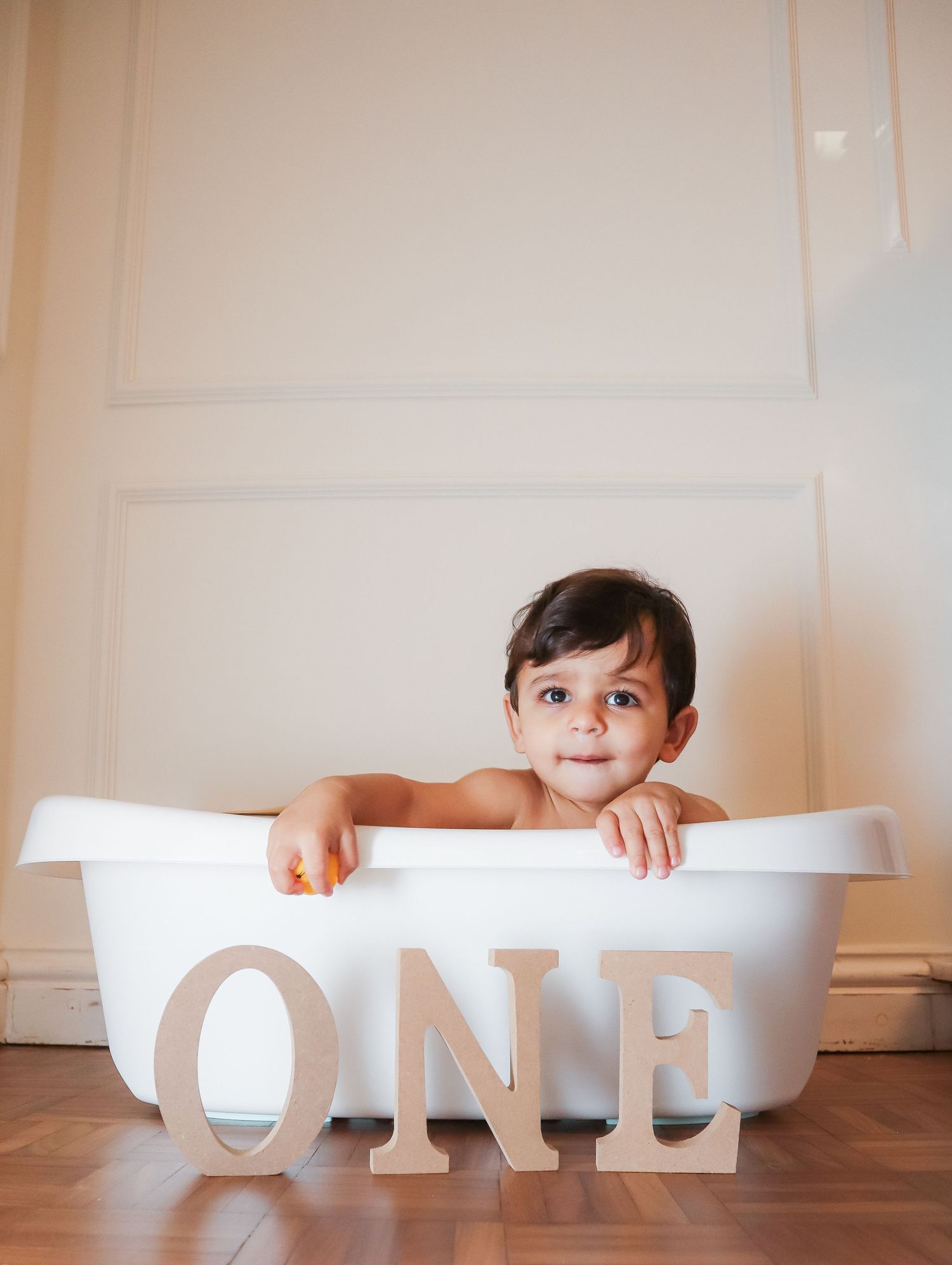 A baby sitting in a white mini bathtub with wooden ONE letters in front, celebrating their first birthday.