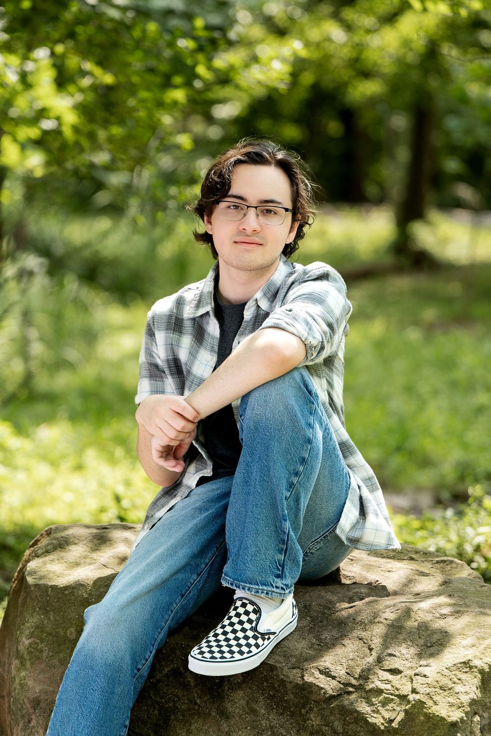 Young man in plaid shirt and checkered Vans sitting on large rock in sunny green park setting.