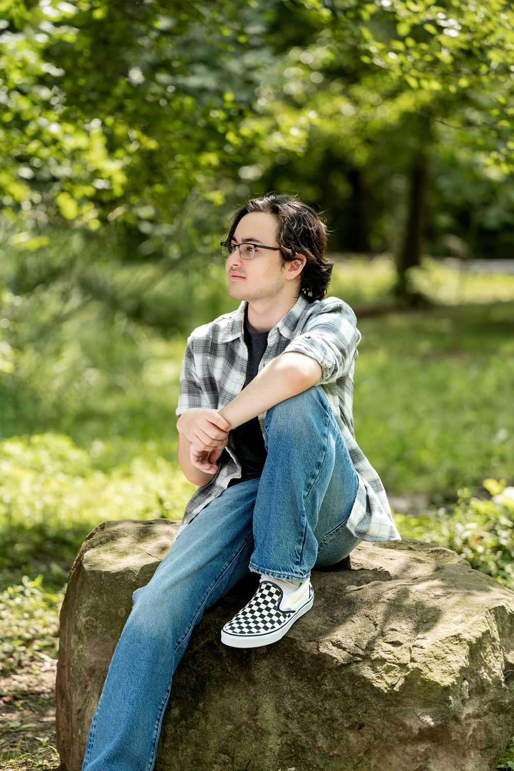 Teenage boy with glasses in plaid shirt sitting on boulder, gazing away in a sunlit green forest.
