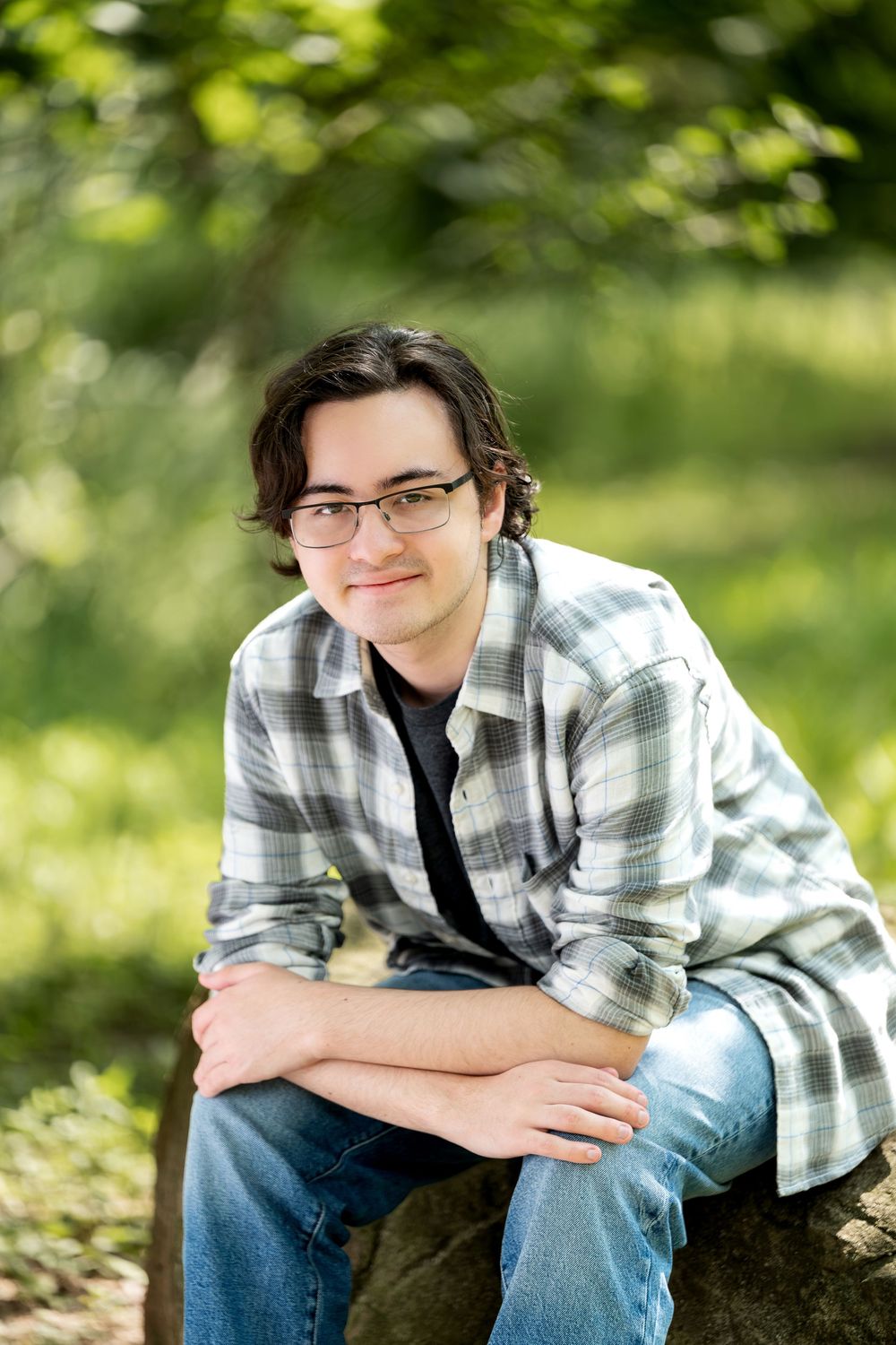 Young man with glasses in plaid shirt smiling outdoors, seated on a rock surrounded by lush green trees.