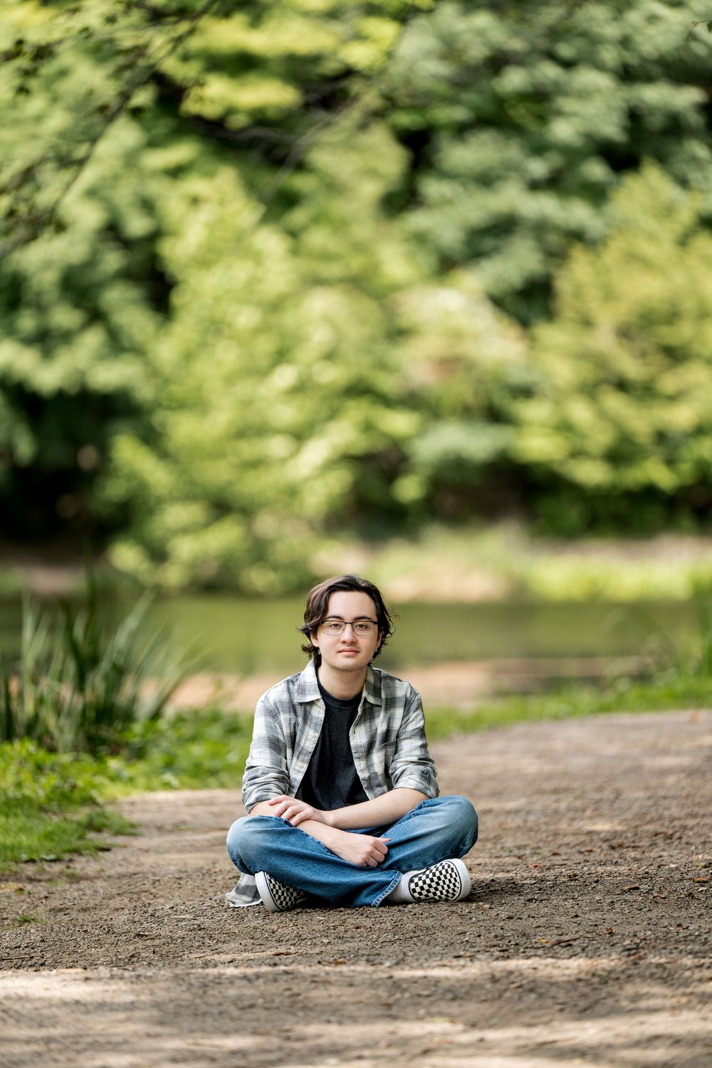 Young man with glasses sitting cross-legged on a park path surrounded by lush green trees near a pond.