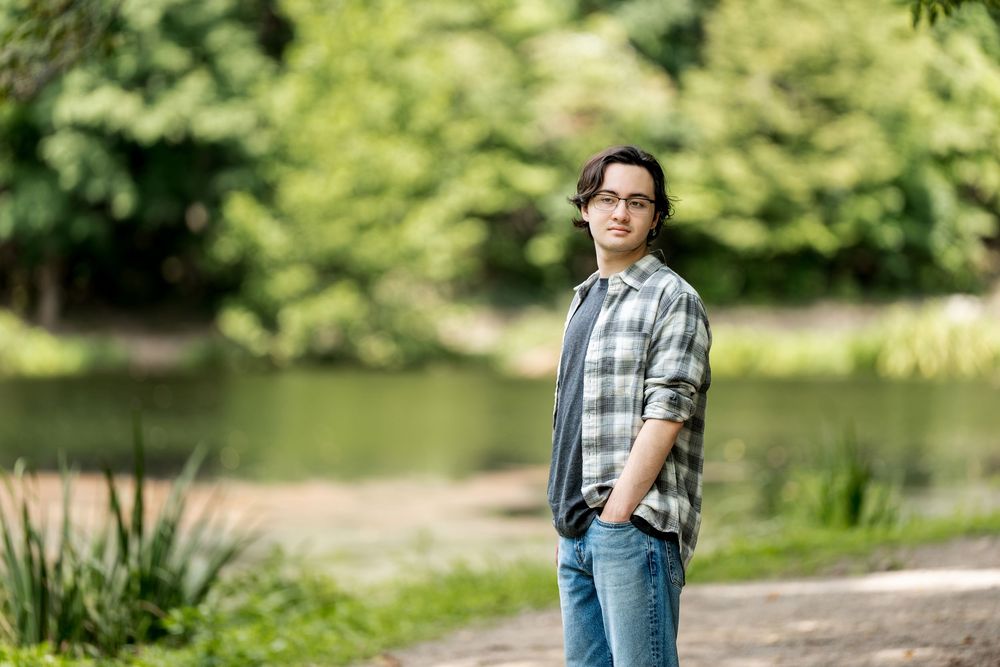 Young man in plaid shirt and jeans standing in a lush green park near a pond.