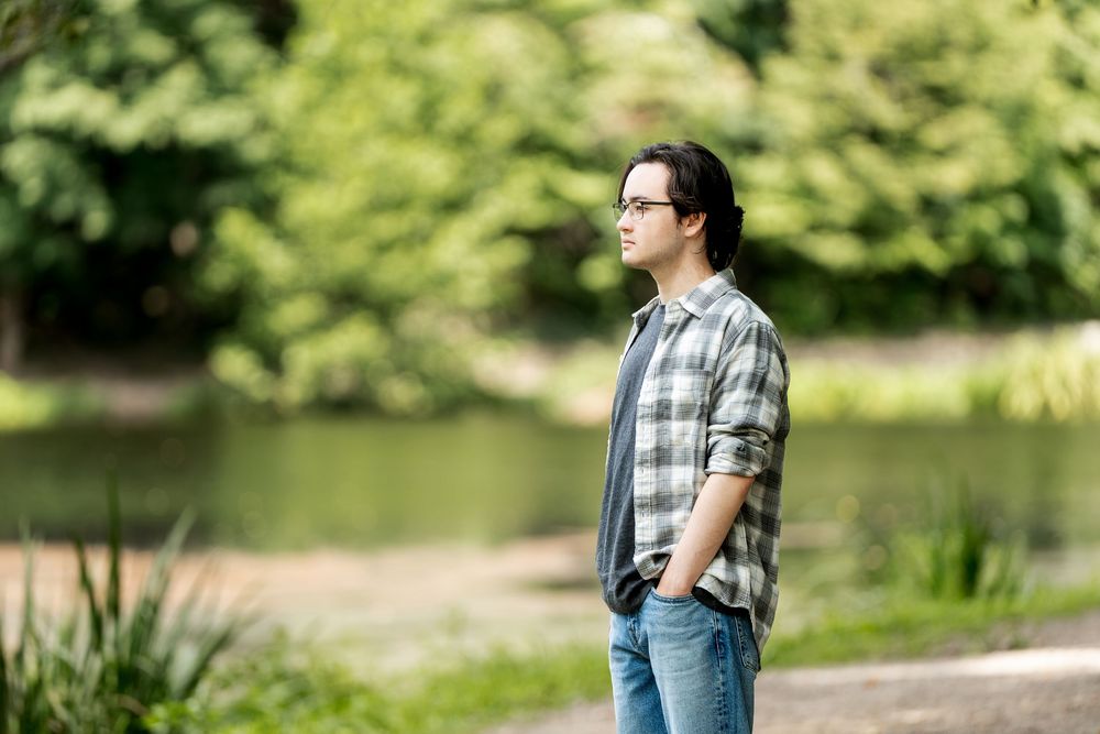 Young man with glasses standing outdoors near a pond, wearing a plaid shirt and jeans in a lush green park.