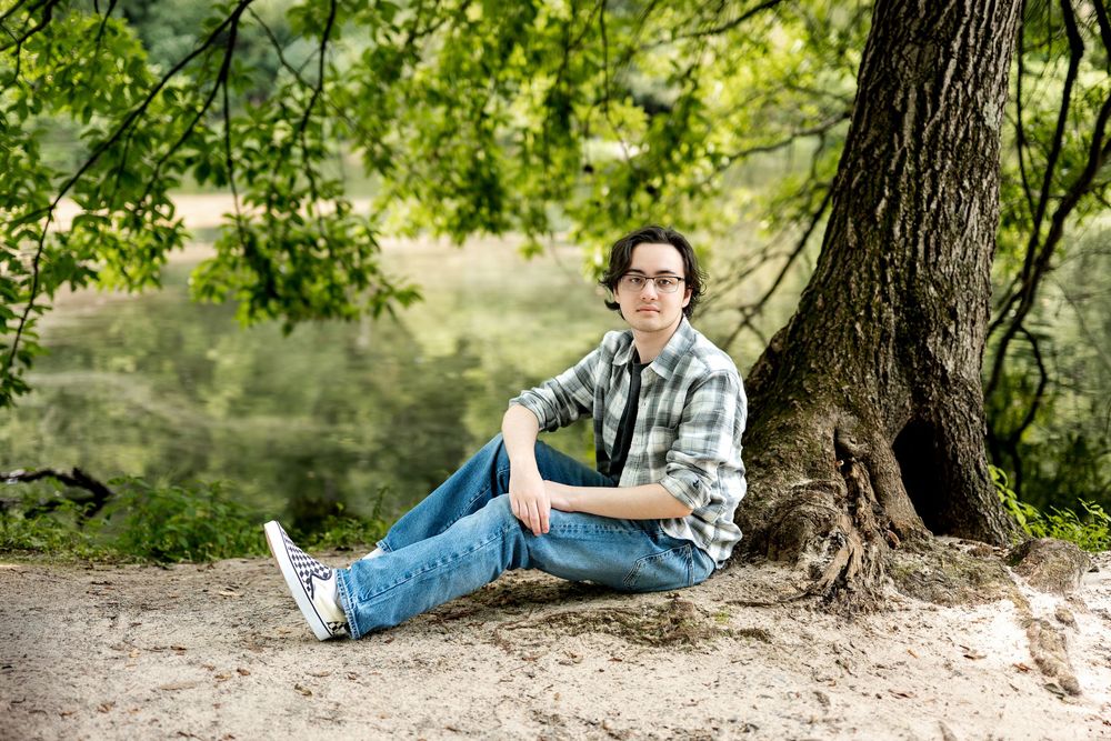 Teen boy with glasses leaning against a large tree trunk in a lush green park setting.
