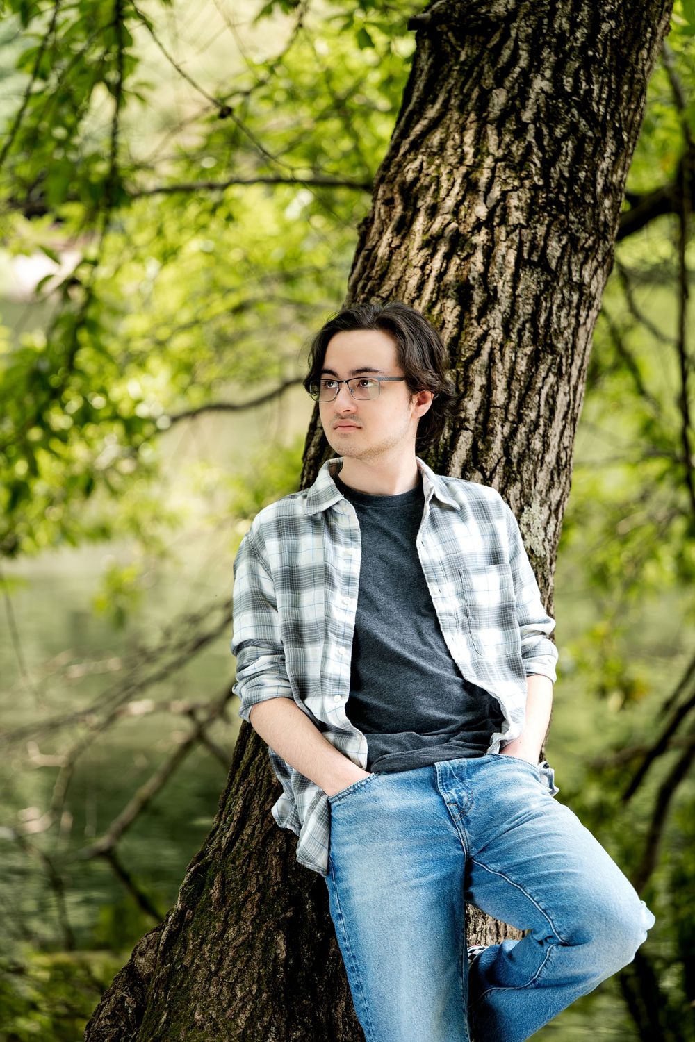 Young man with glasses in plaid shirt leaning against a tree trunk outdoors near a tranquil green forest.