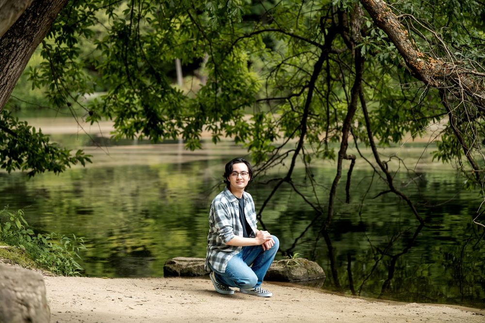 Smiling young man with glasses crouching near a tranquil pond under overhanging tree branches in a park.