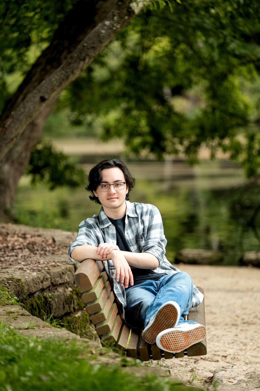 Young man with glasses sitting on a stone wall in a lush green park, wearing a plaid shirt and jeans.