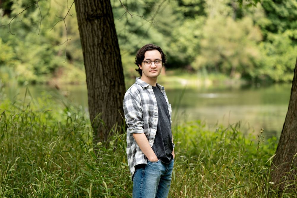 Young man in plaid shirt and jeans standing among trees near a pond in a lush green forest setting.
