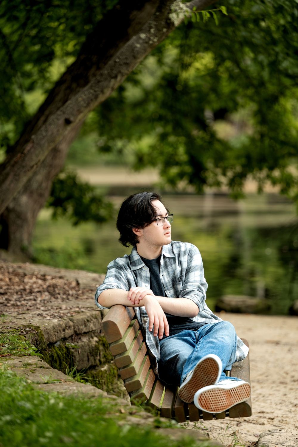 Teen boy in plaid shirt and jeans sits by a pond under green trees, gazing thoughtfully into the distance.