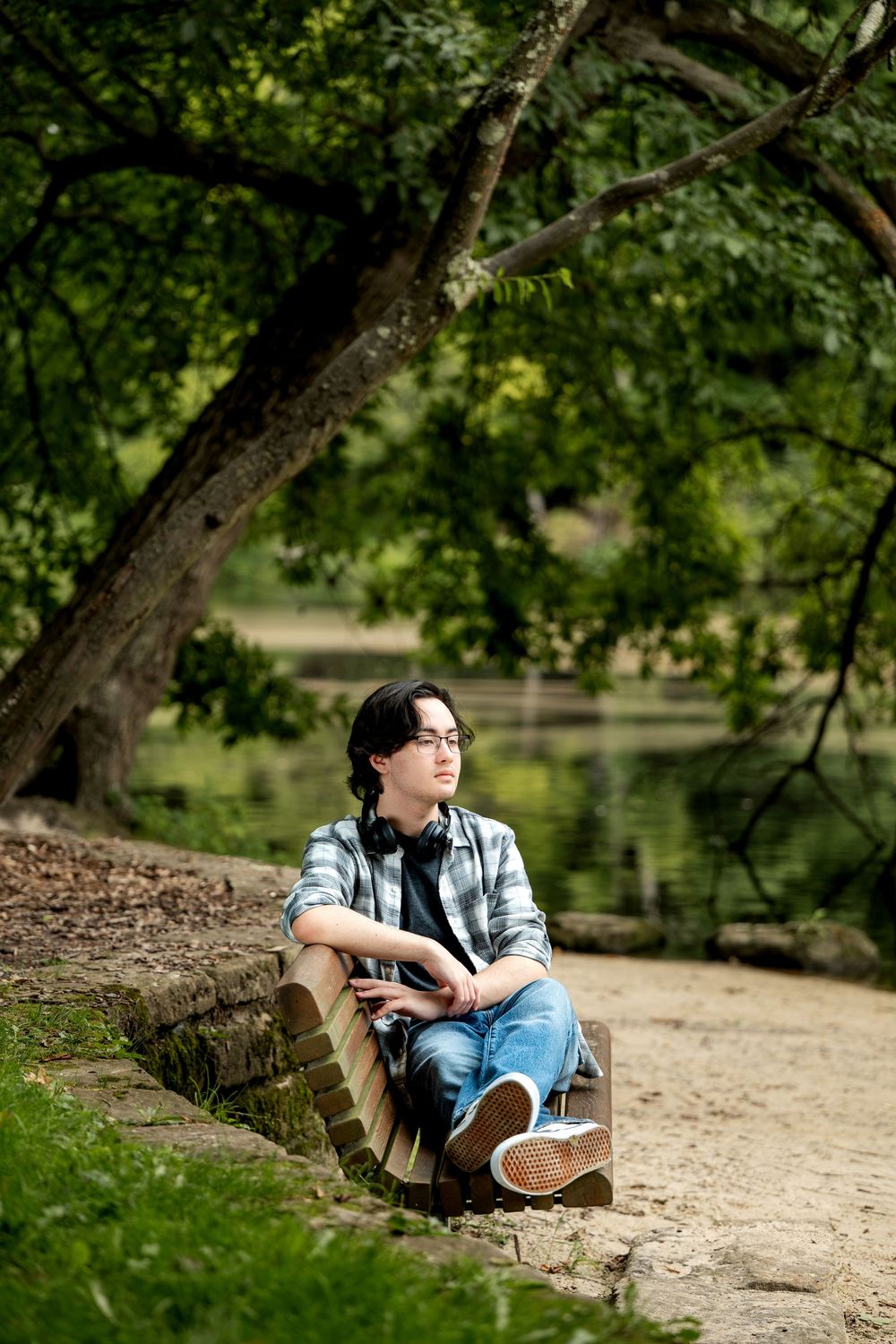 Young person in plaid shirt and jeans sitting on a bench in a lush green park near a pond.