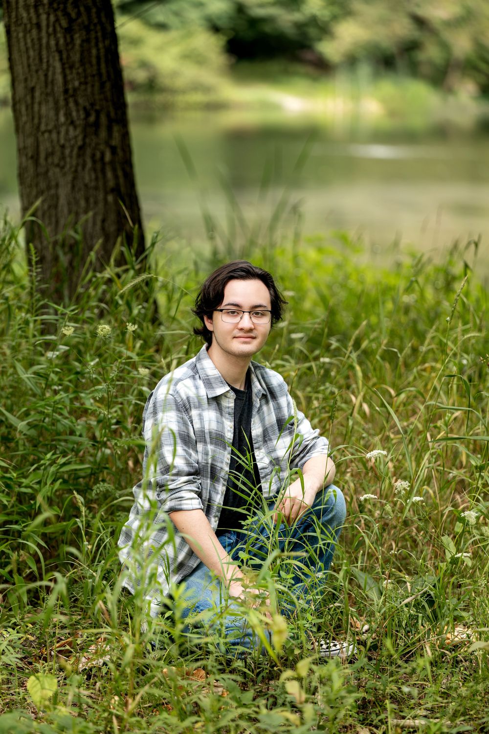 Young man with glasses sitting in tall grass near a peaceful pond surrounded by lush greenery.