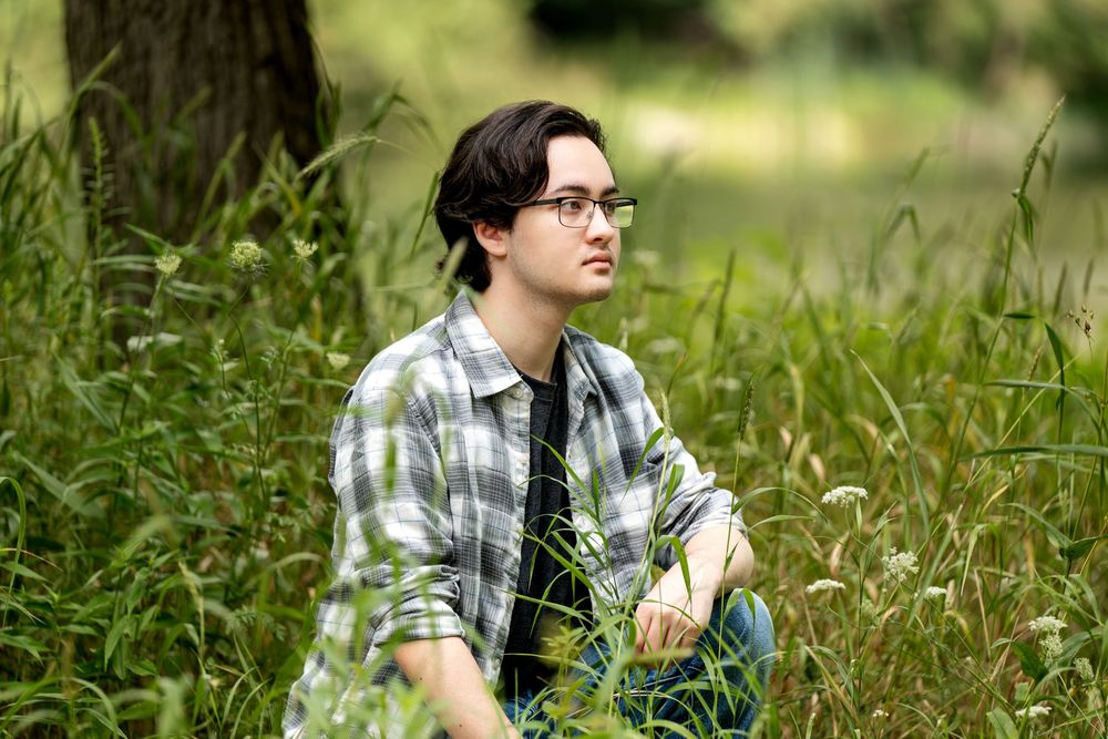 Young man with glasses sitting in tall green grass outdoors, wearing a plaid shirt, looking thoughtfully to the side.