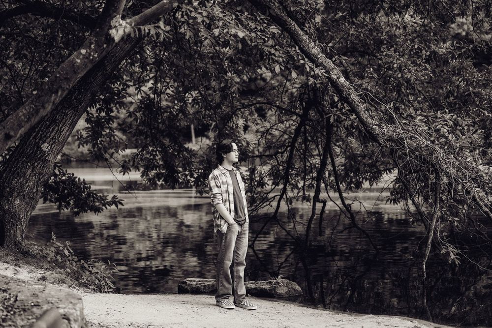 Black and white portrait of a young man standing under a large tree beside a reflective pond.