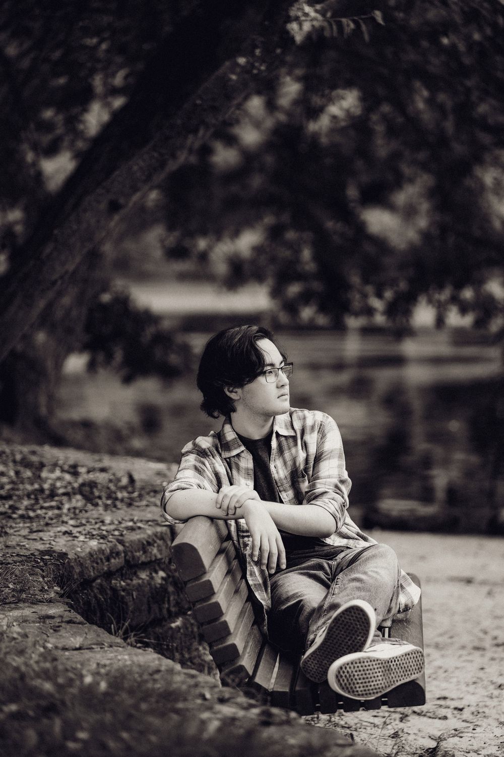 Black and white portrait of a young man sitting by a tree near a pond in a serene outdoor setting.