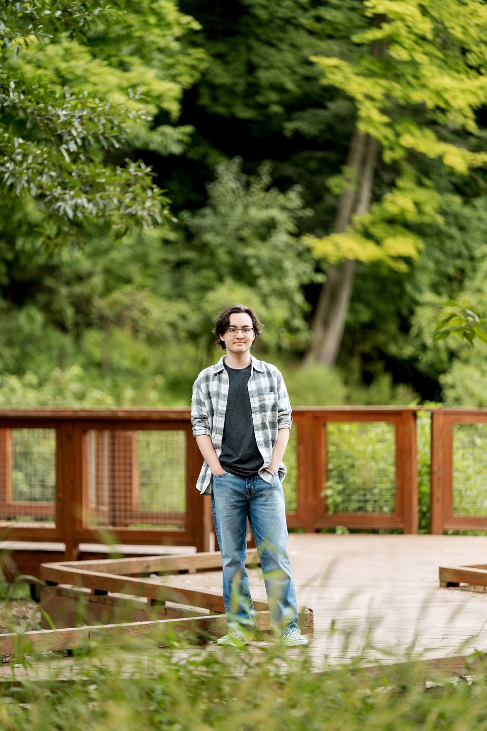 Young man in plaid shirt and jeans smiling on a wooden bridge surrounded by lush green trees.