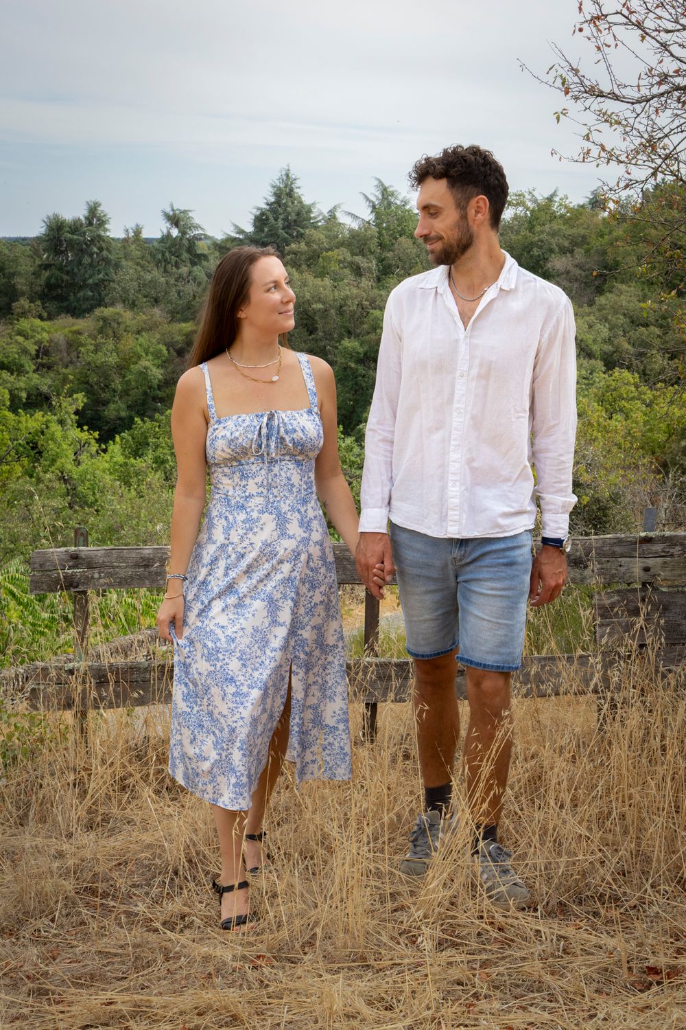 Couple de fiancés debout dans un champ d’herbes sèches, elle en robe fleurie bleue, lui en chemise blanche, Angers