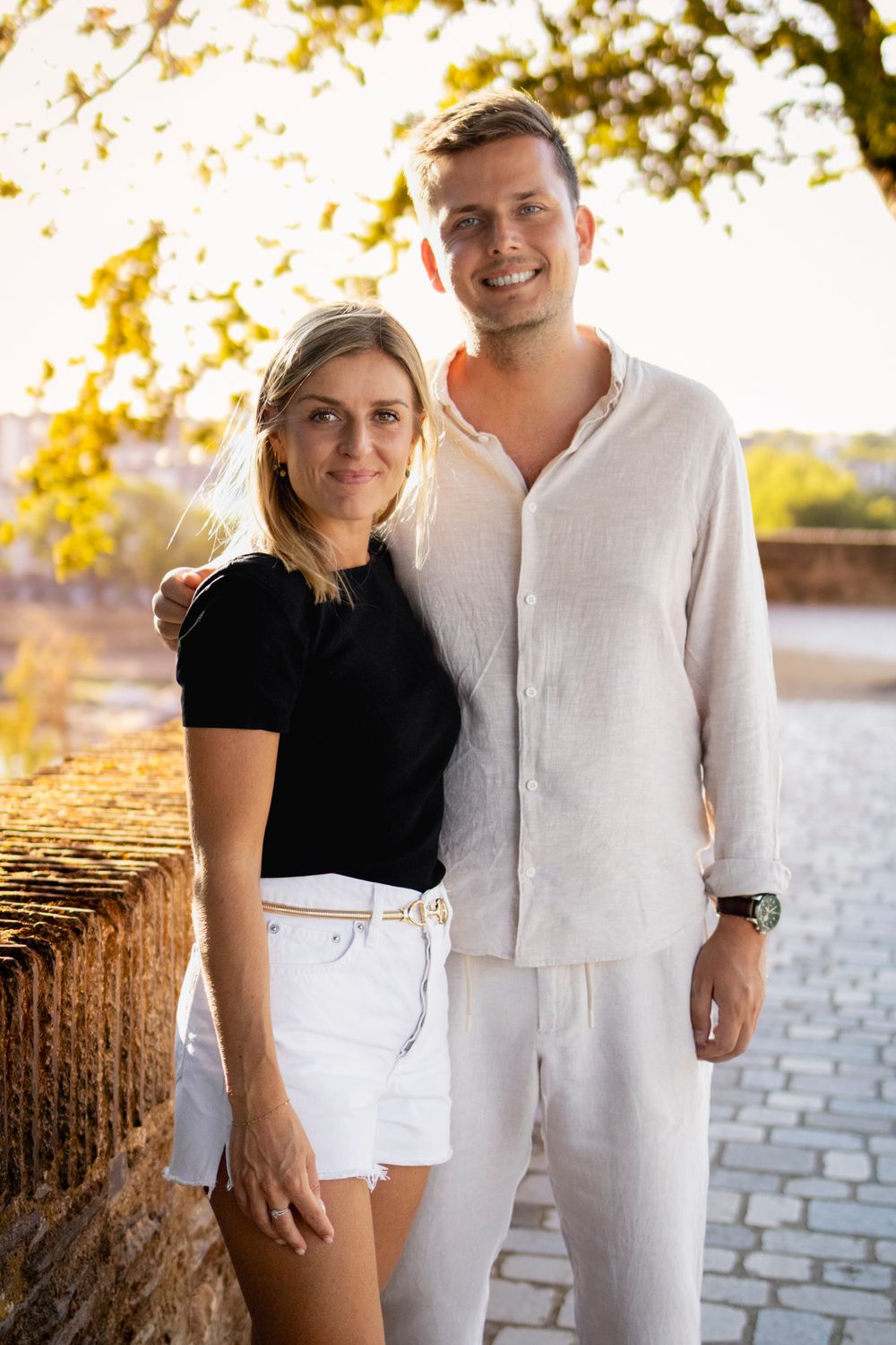 Couple souriant debout sur des pavés, feuillage doré en arrière-plan, séance photo à Angers