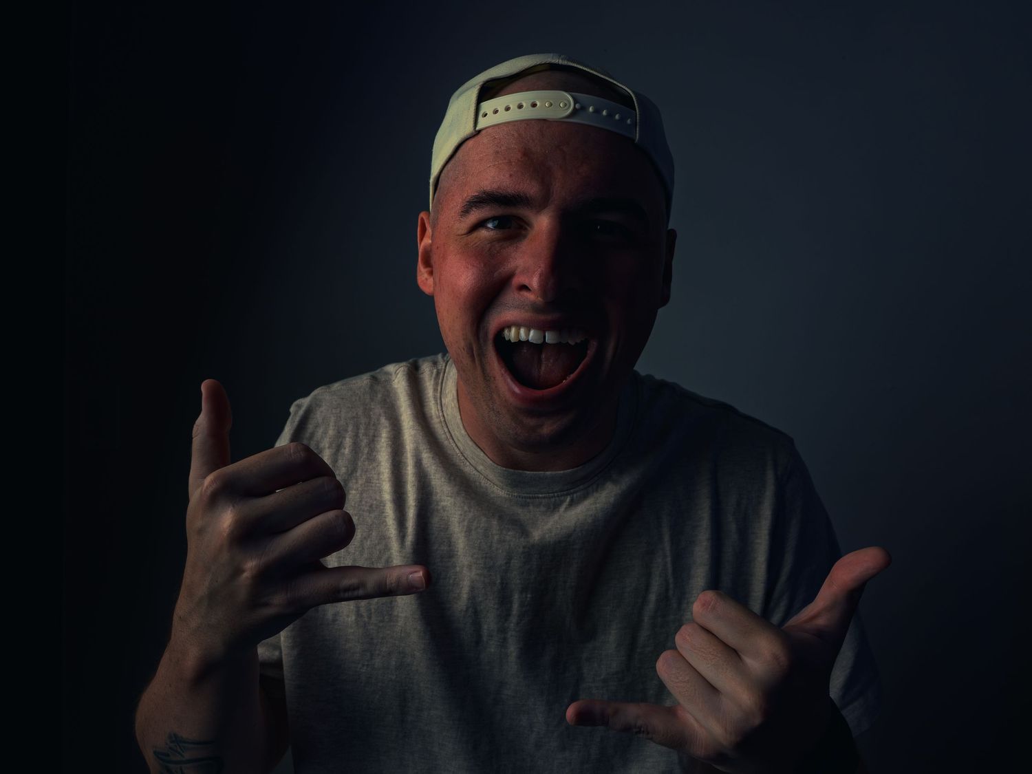 A man in a backward cap and gray tee laughs wide, throwing double shaka signs against a dark moody backdrop, a forearm tattoo showing