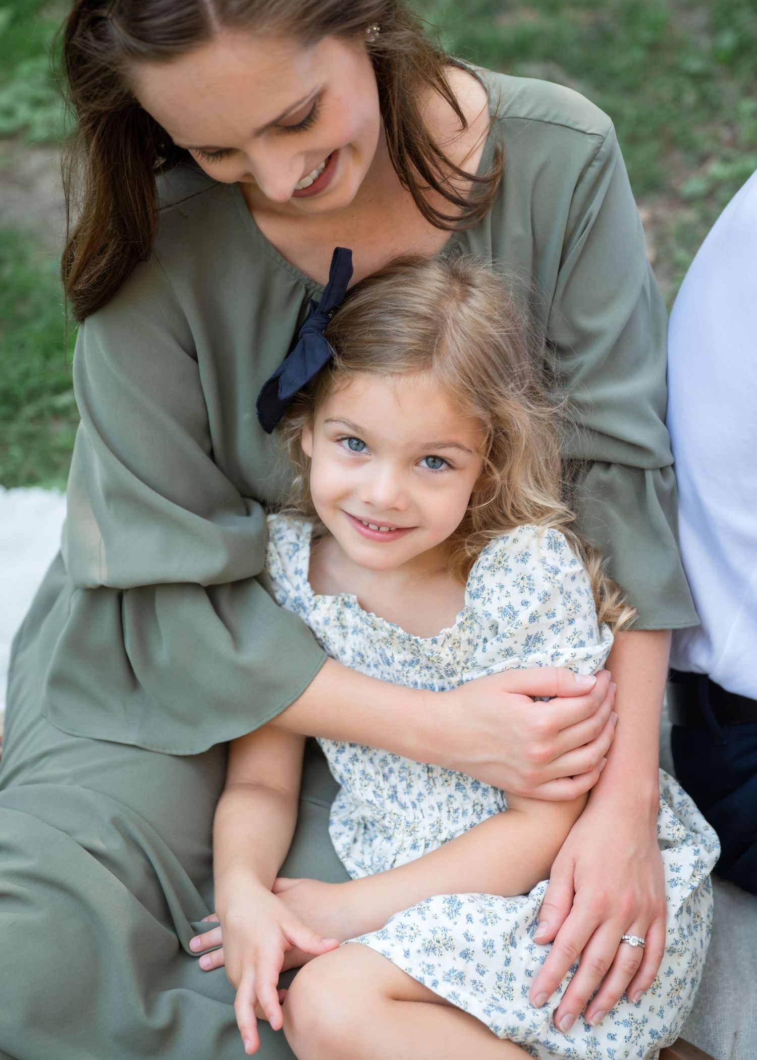 A woman in sage green dress smiles while hugging a young girl in floral dress during an outdoor family photo session.