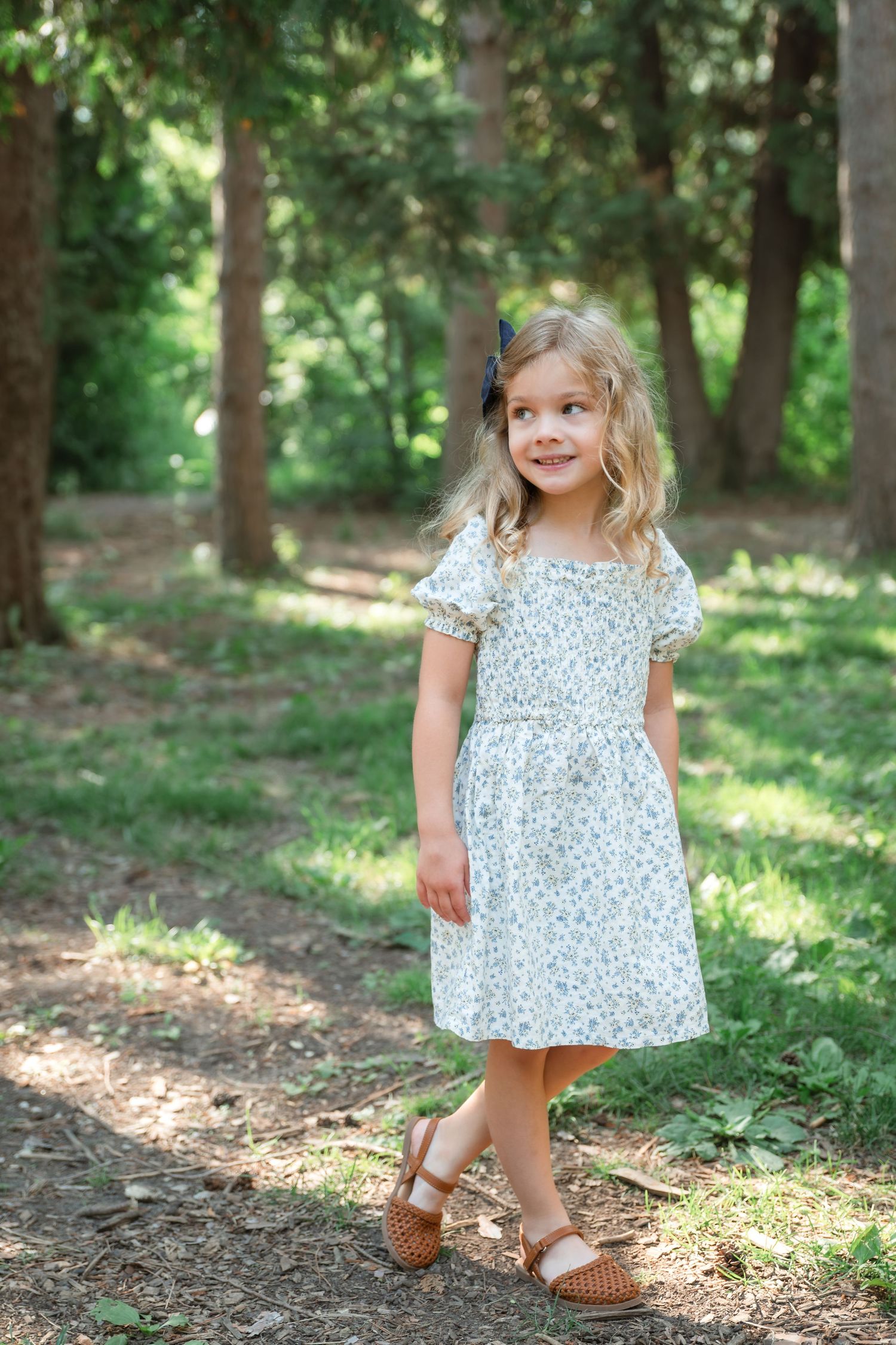 A smiling young girl with blonde curly hair and a navy bow poses beside a tree in a floral dress outdoors.