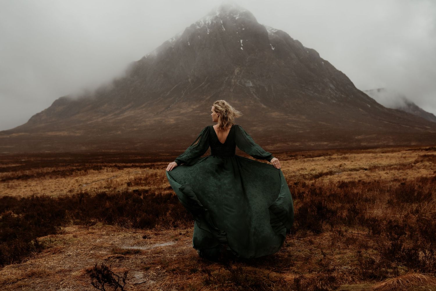 Woman in flowing green dress twirls before misty mountain landscape in dramatic Scottish Highlands scenery.