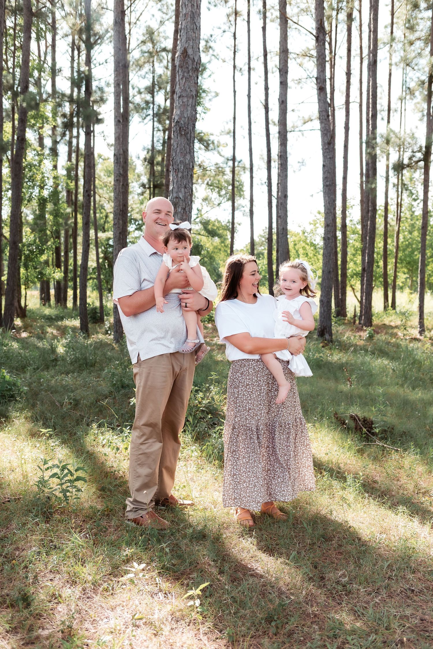 Family of four enjoying a sunny outdoor photoshoot in a pine forest, parents holding two young daughters.