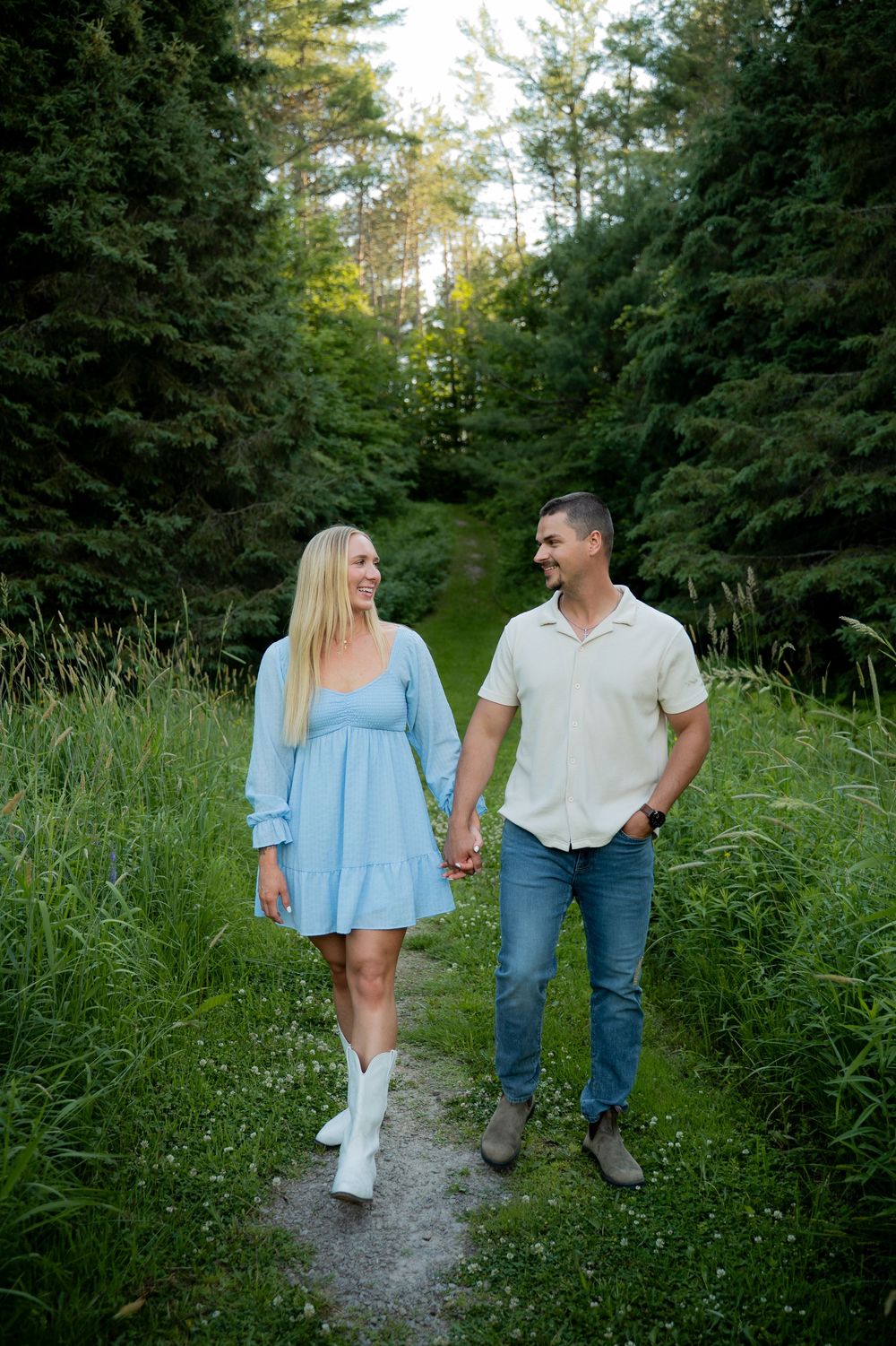 Couple holding hands walking on a forest path, woman in blue dress and white boots, man in white shirt and jeans.