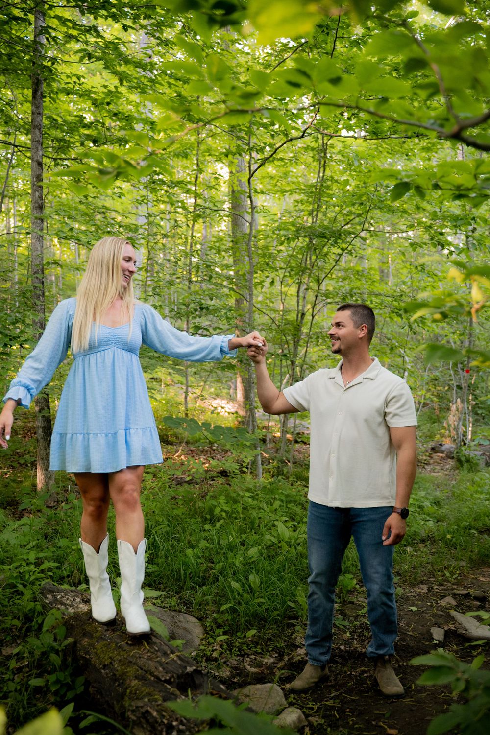 Couple holding hands in a lush green forest, woman in blue dress and white cowboy boots, man in white shirt and jeans.