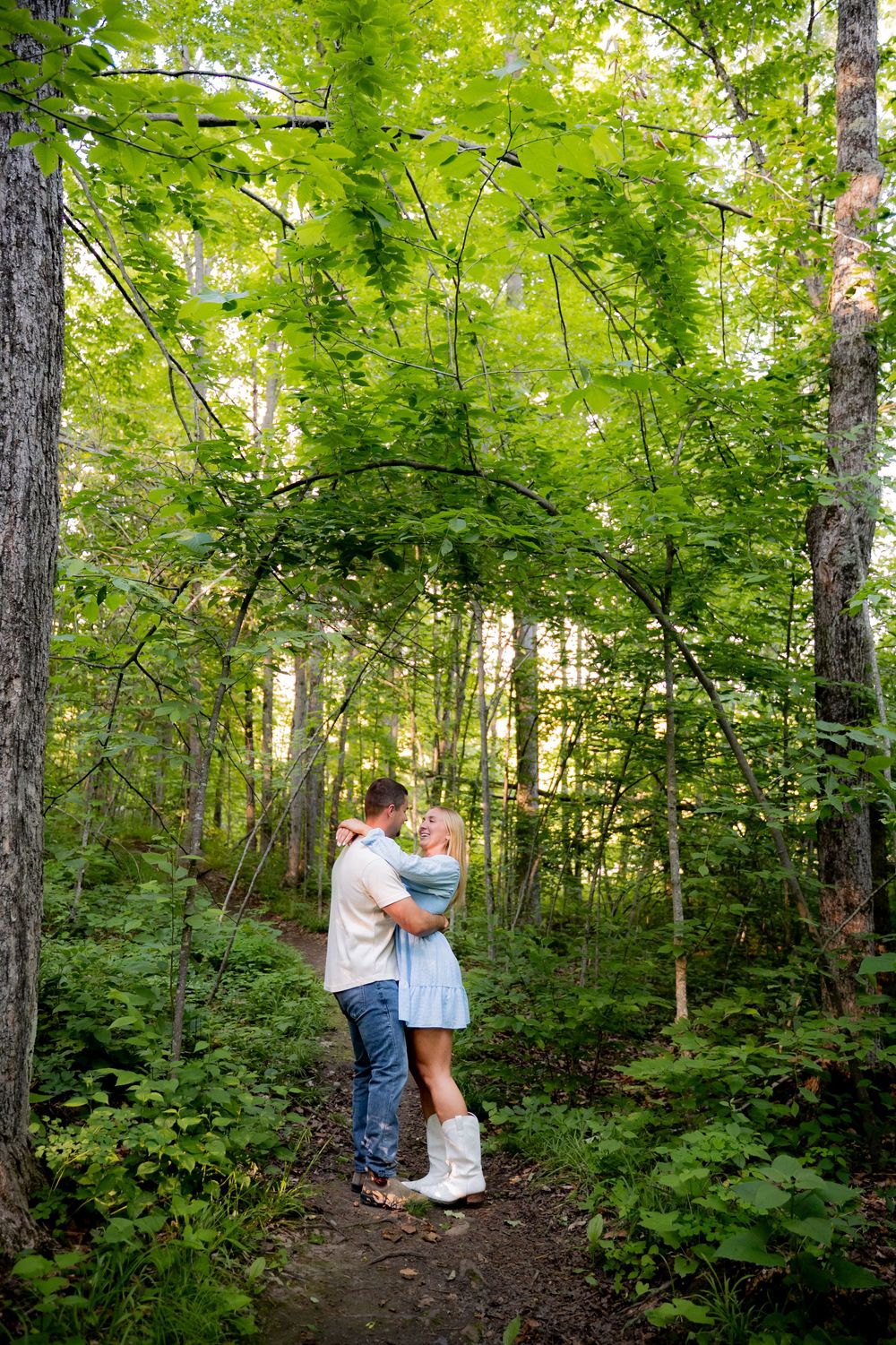 Couple embracing on a forest path surrounded by lush green trees and dappled sunlight.