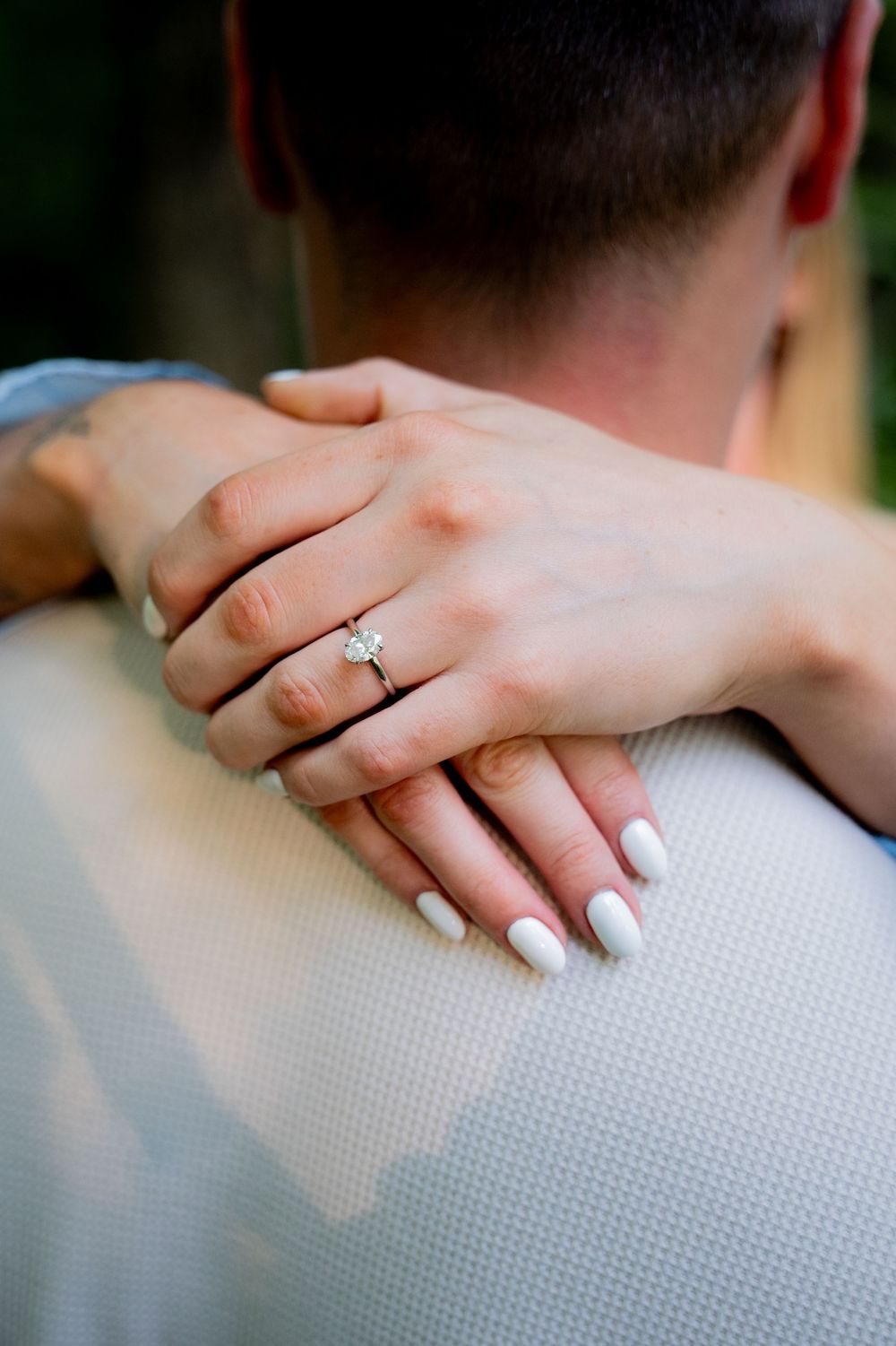 Close-up of a woman's hand with white nails and diamond engagement ring resting on a man's shoulder.