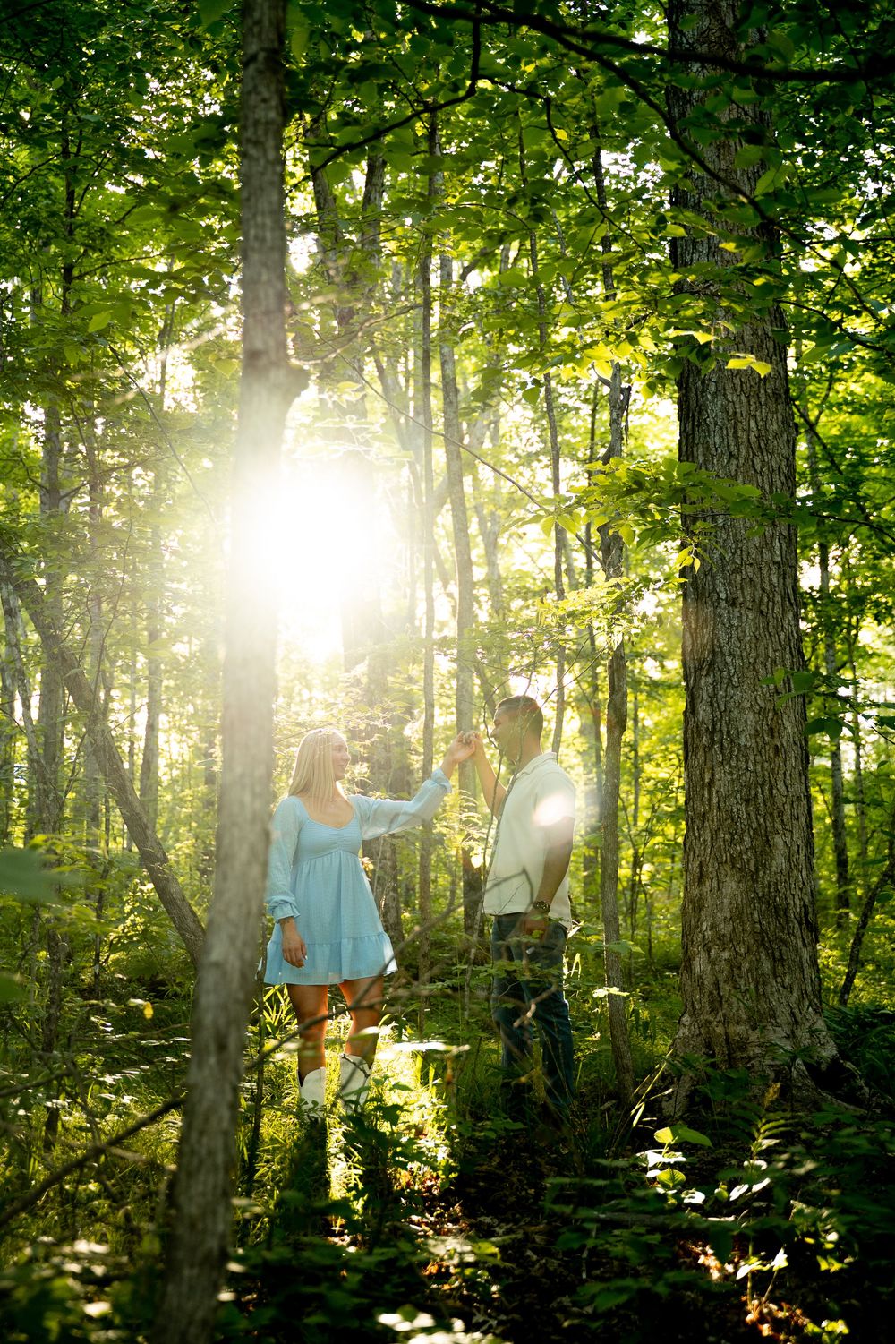 Couple standing in sunlit forest, woman in blue dress and man in white shirt, surrounded by lush green trees.
