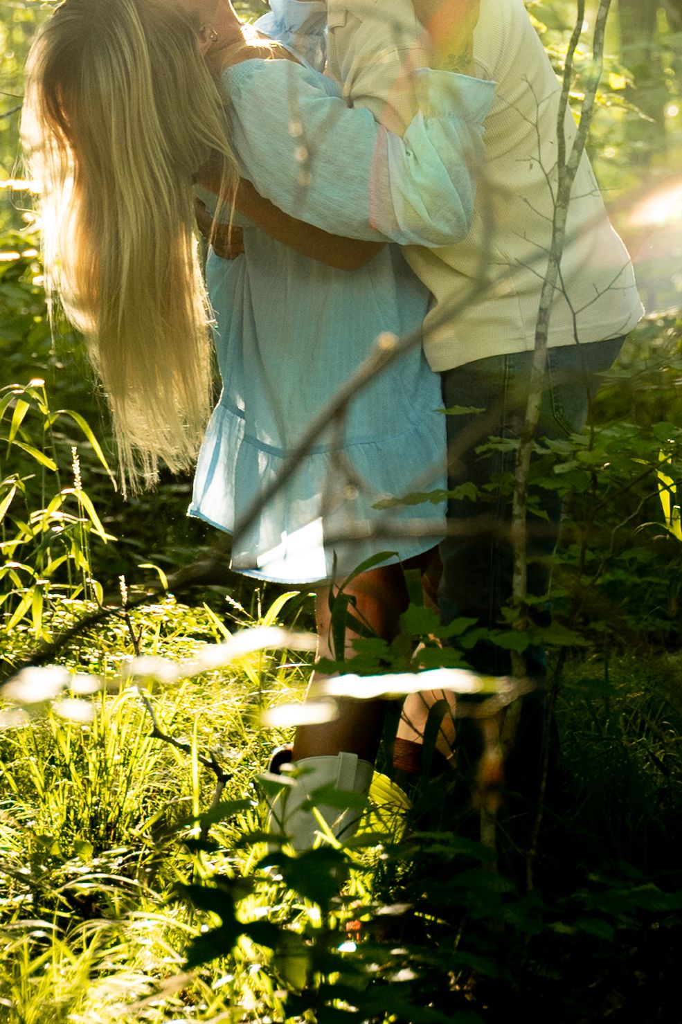 Couple embracing in sunlit forest clearing, surrounded by lush green plants and dappled light.