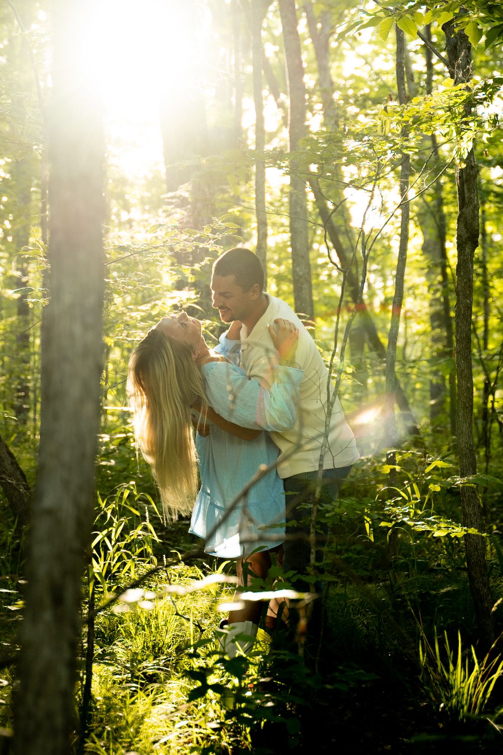 Romantic couple kissing in a sunlit forest with golden light streaming through green trees.