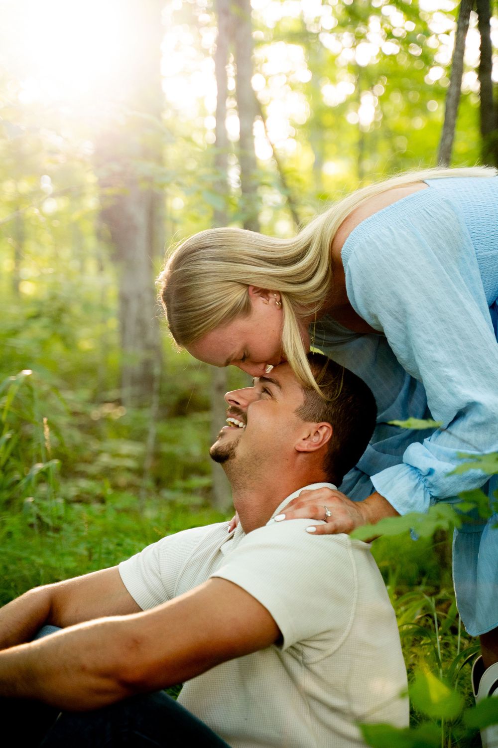 Happy couple sharing a romantic moment in a sunlit forest, woman leaning over smiling man sitting on grass.