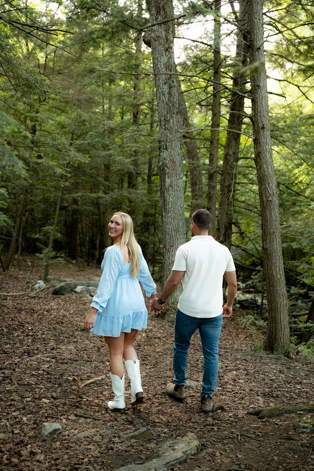 Couple holding hands walking on a forest trail, woman in blue dress and white boots, man in white shirt and jeans.